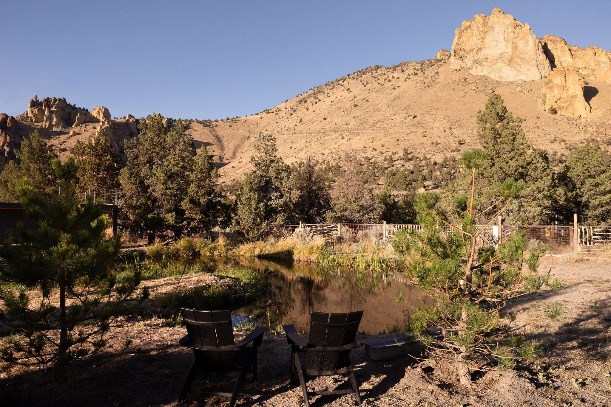 A desert landscape with mountains, trees, and a small pond with two black chairs facing it.