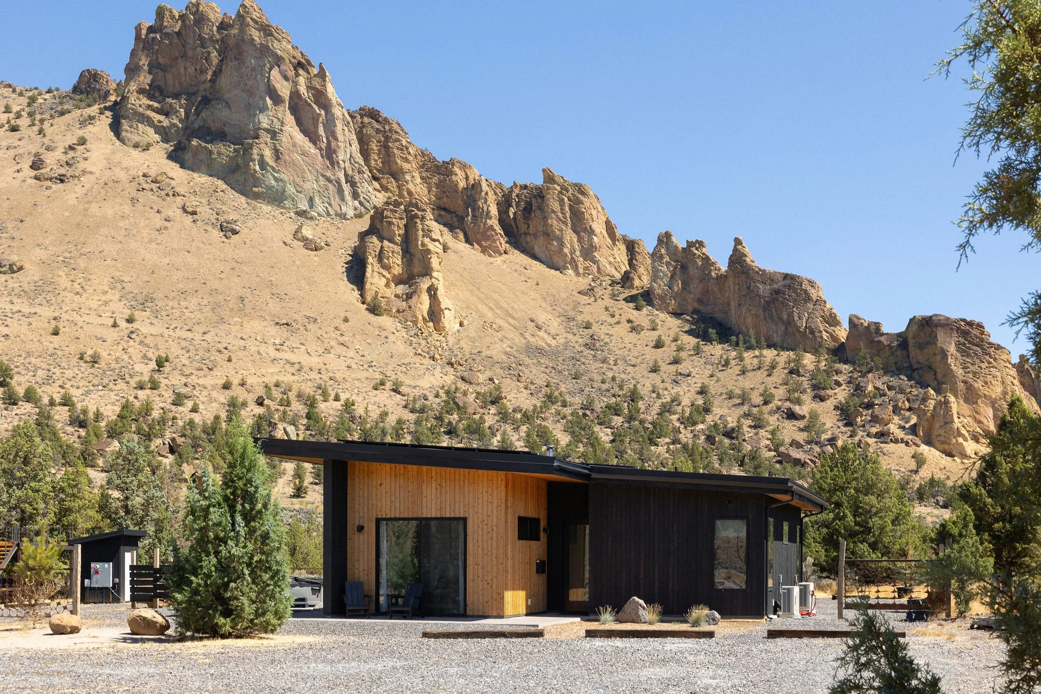 A modern house with a black and wooden exterior, situated in a desert landscape with mountains in the background, trees, and a clear blue sky.