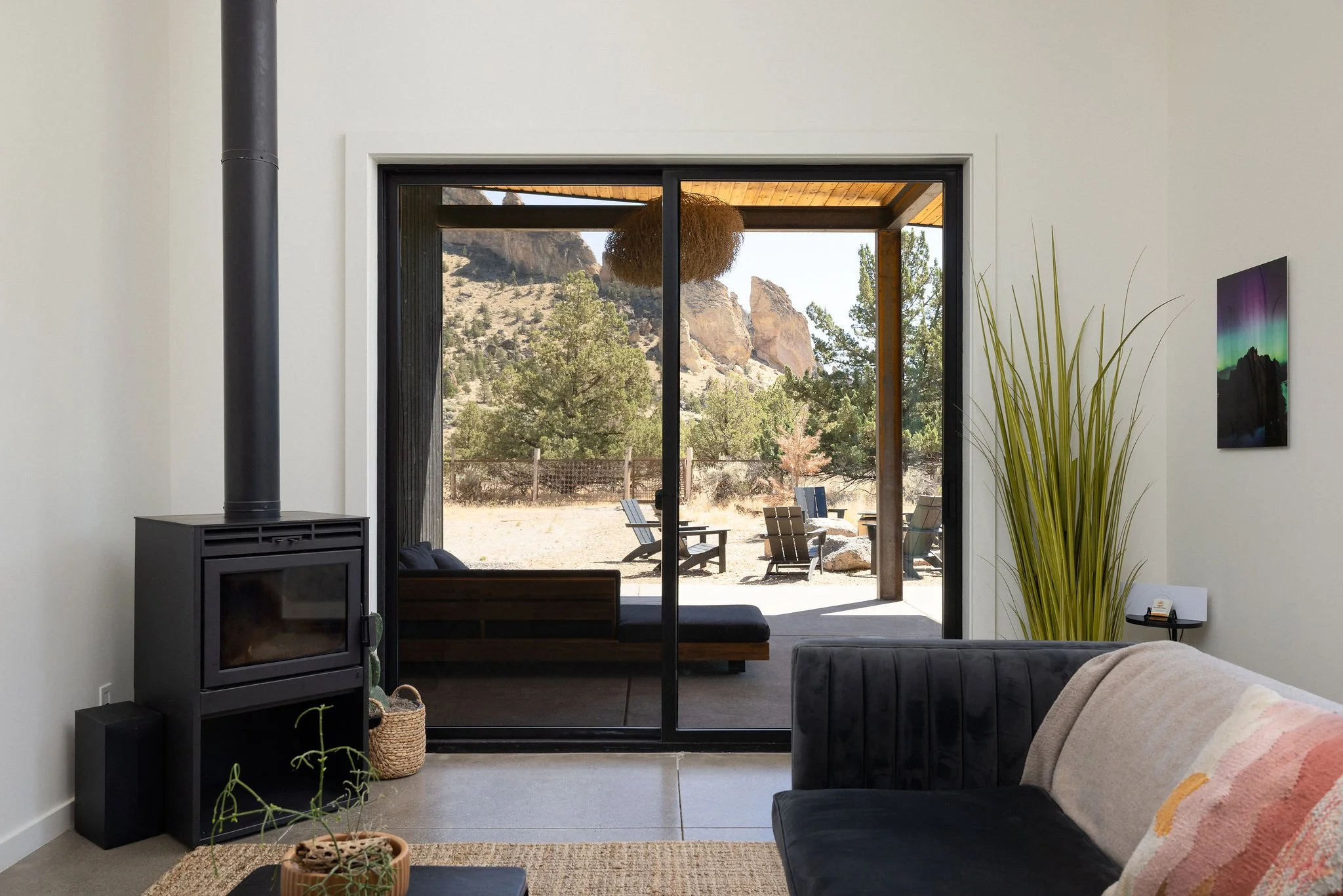 Living room with sliding glass door leading to outdoor patio with chairs and trees; interior features a black wood stove, green plant, framed artwork, and a gray sofa with a beige and pink throw blanket.