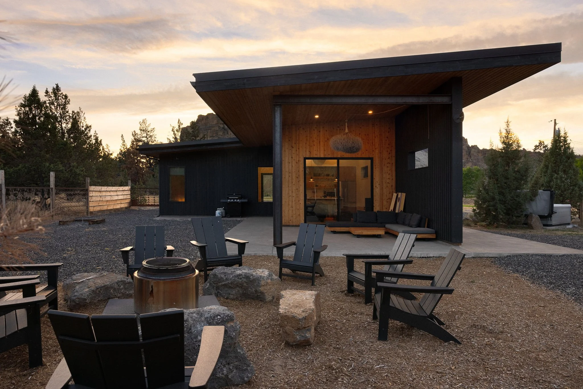 Modern house with an outdoor patio area featuring black Adirondack chairs around a fire pit, set against a natural landscape at sunset.