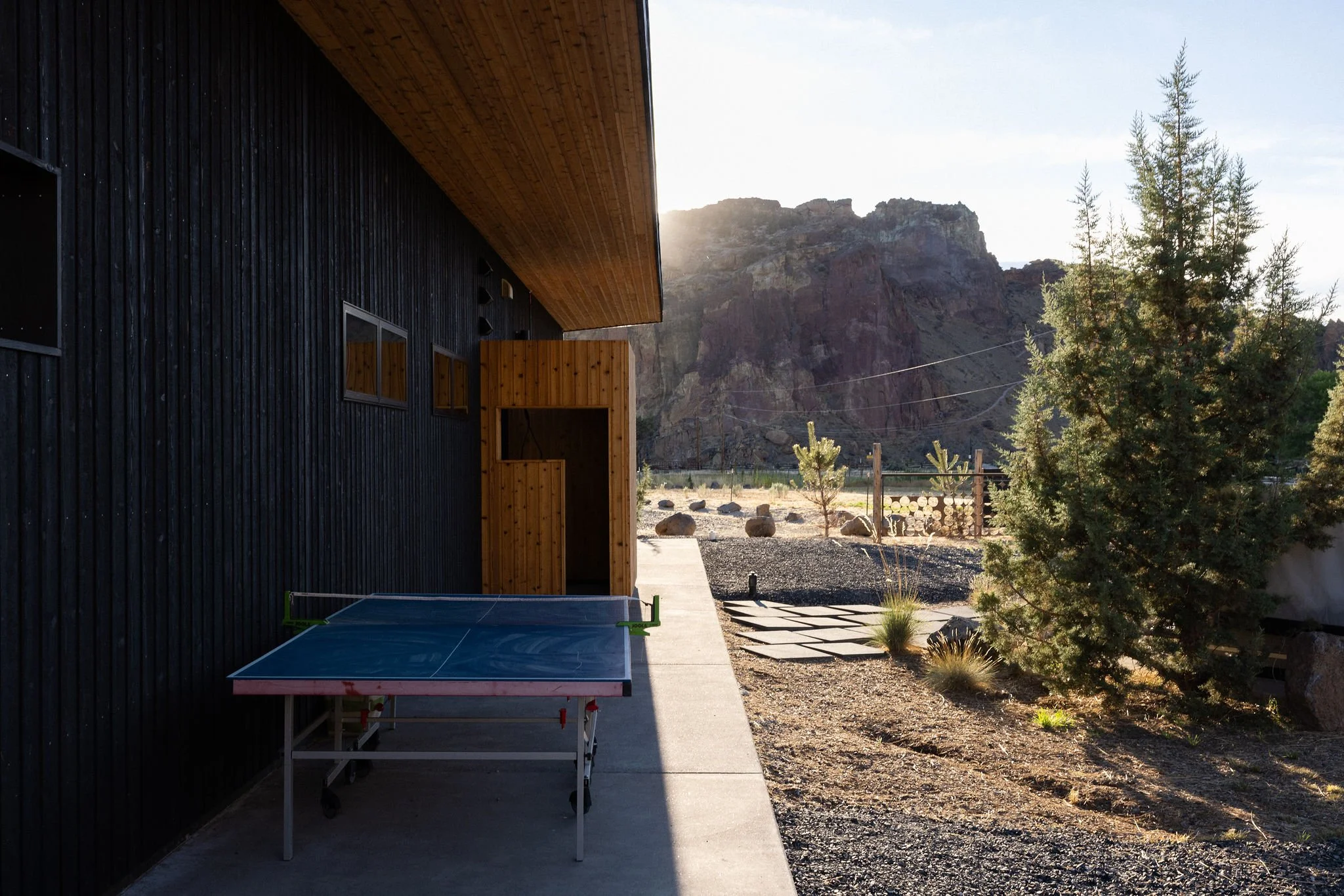 View of a modern house with black and wood exterior, a ping pong table outside, and a mountainous desert landscape in the background.