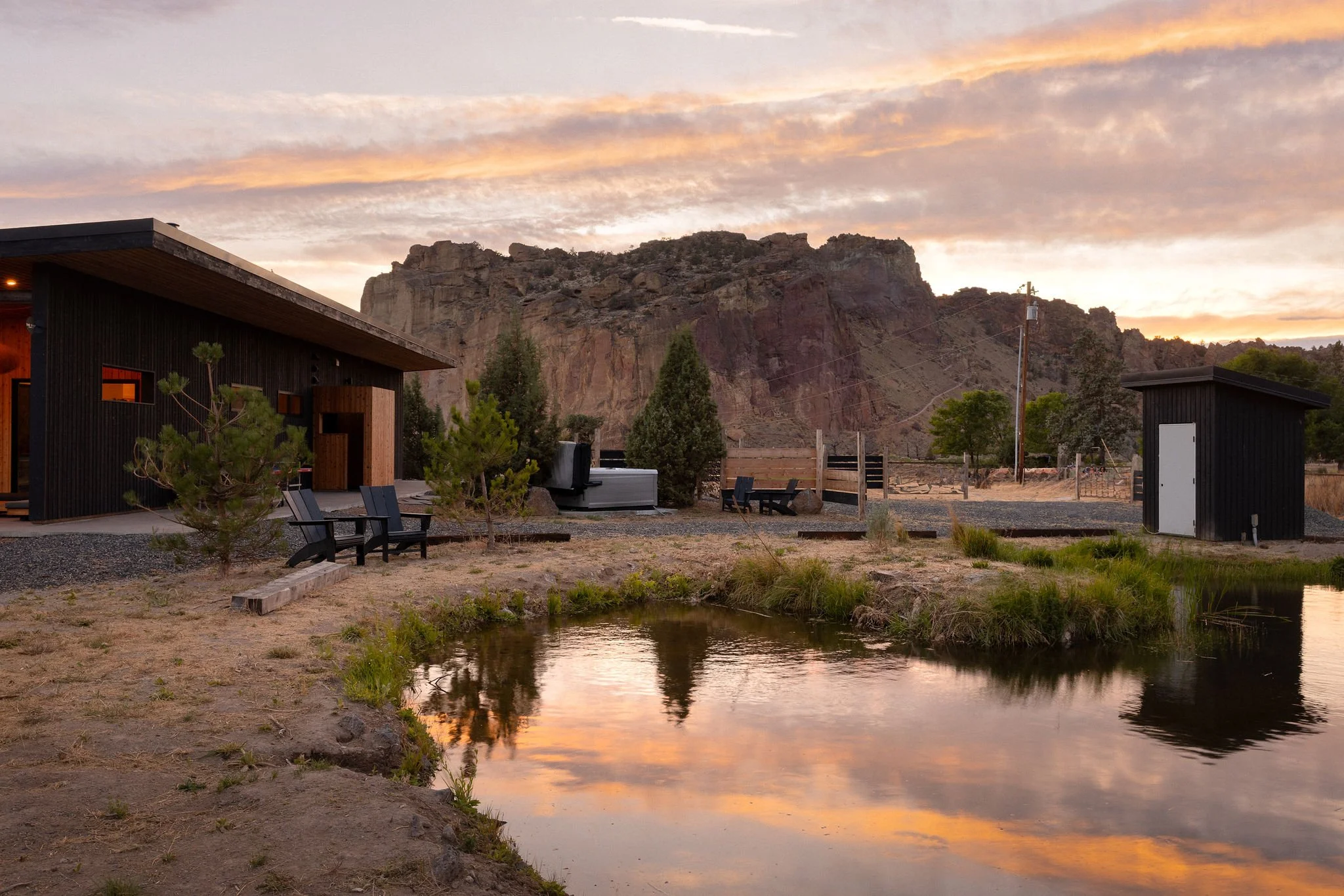 A scenic outdoor scene at sunset with modern black buildings, small trees, a pond reflecting the sky, and a mountain in the background.