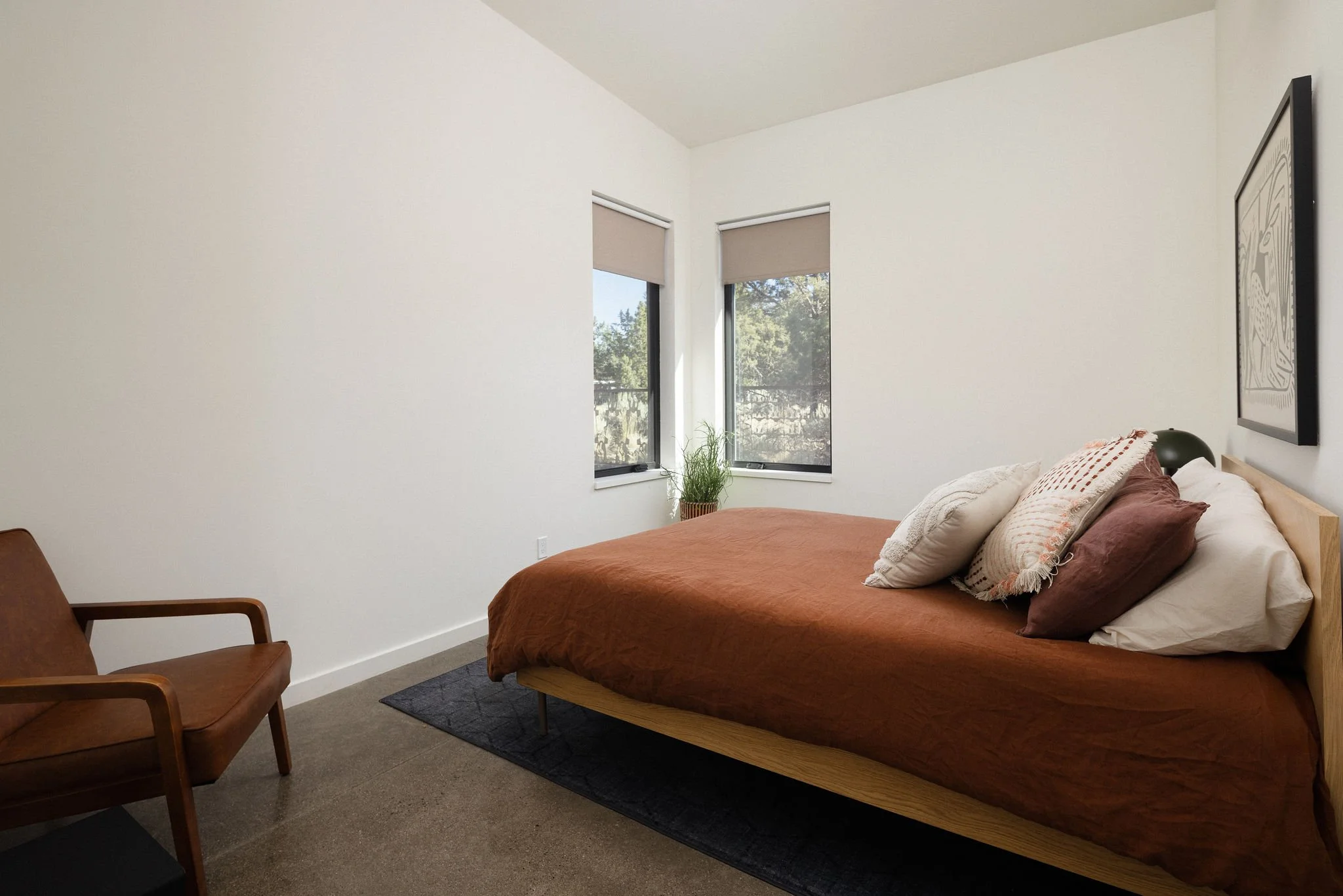 A minimalist bedroom with a wooden bed, brown bedding, multiple pillows, a small black rug, a brown chair, a green potted plant, and two windows with beige blinds.