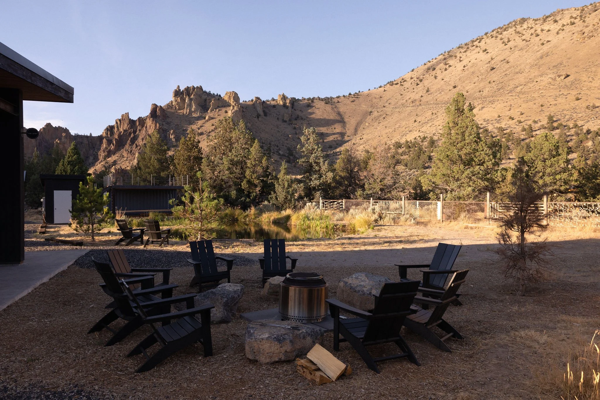An outdoor seating area with six black Adirondack chairs arranged in a circle around a fire pit, with rocks and a small piece of firewood nearby. The area is next to a small pond with green plants, surrounded by trees and a wooden fence, with rocky h