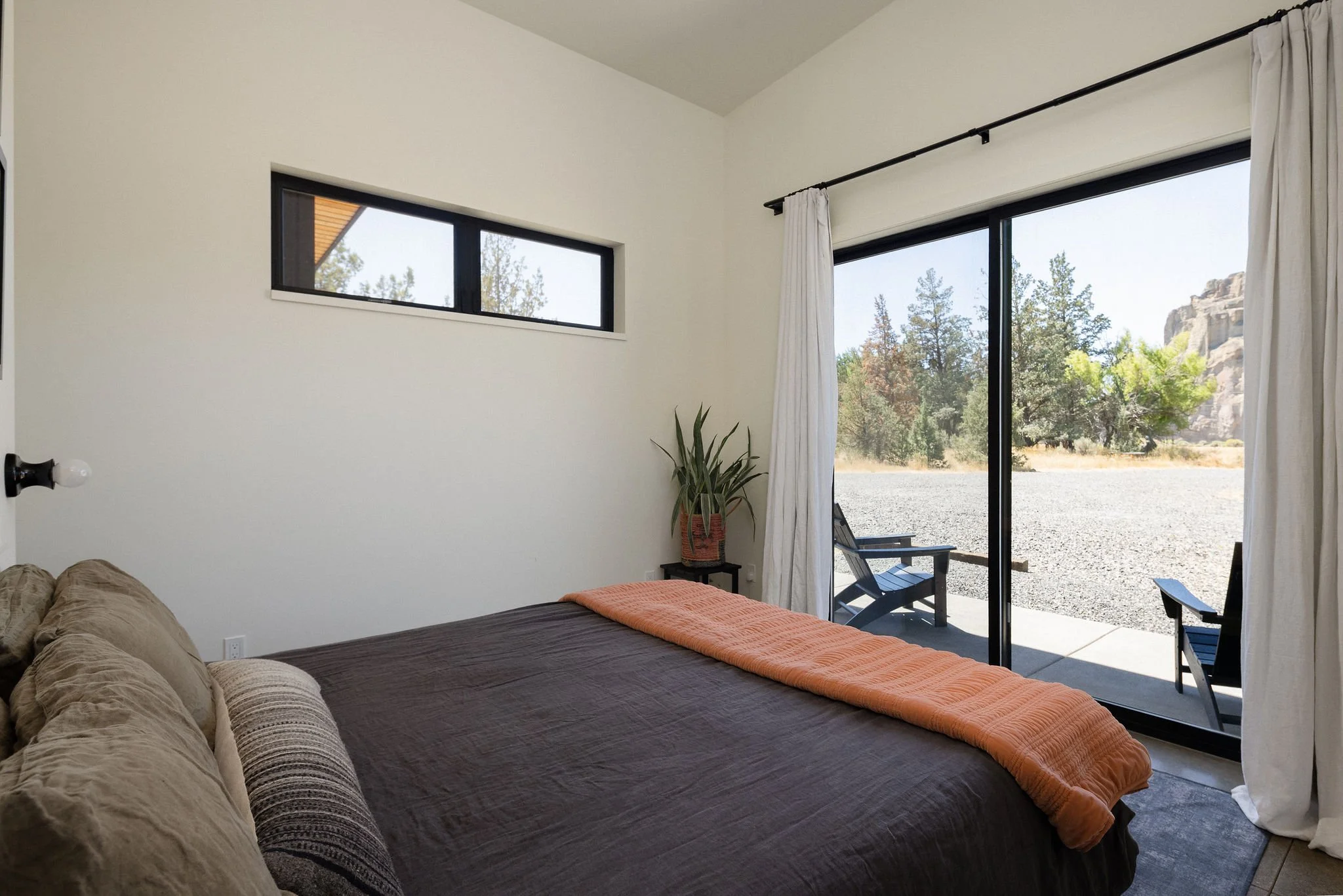 Bedroom with a large sliding glass door showing outdoor chairs and trees, with white curtains and a potted plant in the corner.