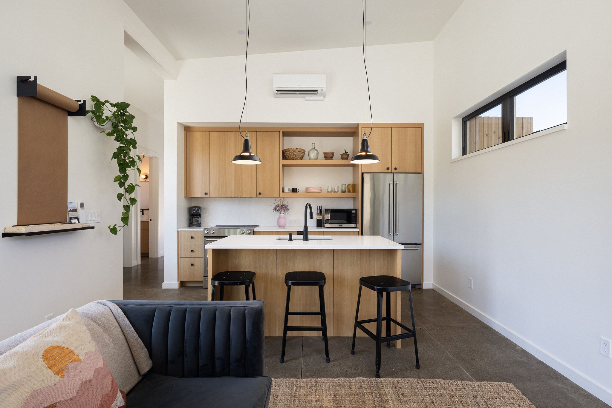 Modern kitchen with light wood cabinets, white island with black faucet, three black stools, stainless steel refrigerator, open shelving, and minimalist decor, next to a living area with a dark sofa and a patterned pillow.