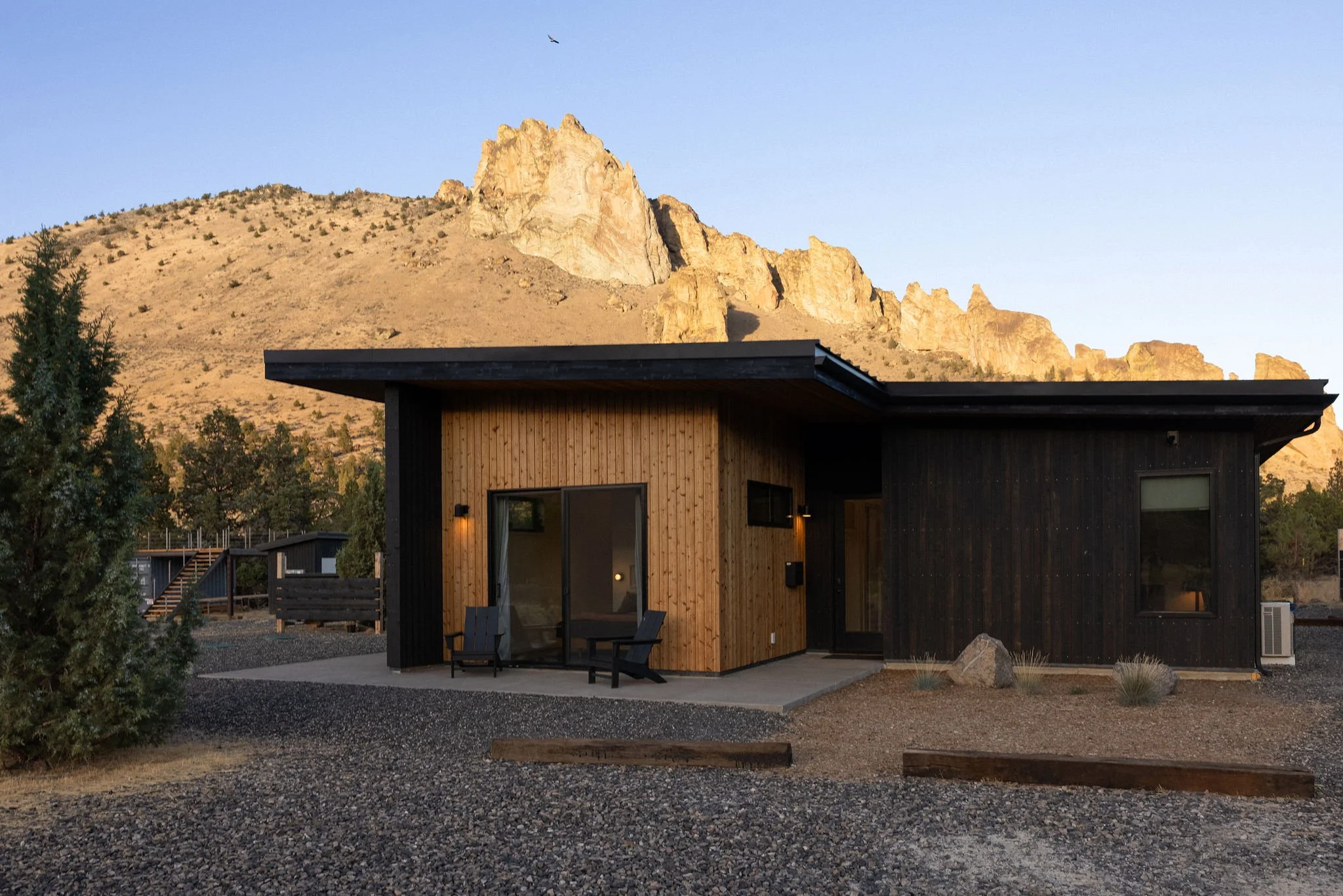 A modern house with wooden and black exterior walls, set against a mountain landscape during sunset