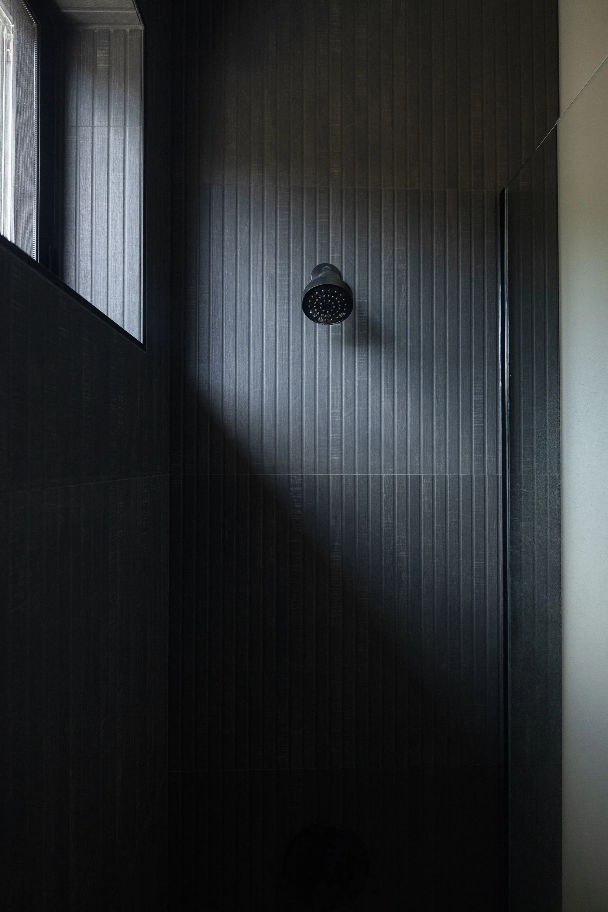 Interior view of a modern shower stall with black textured tiles on the walls, a black showerhead, and a small window on the left side.