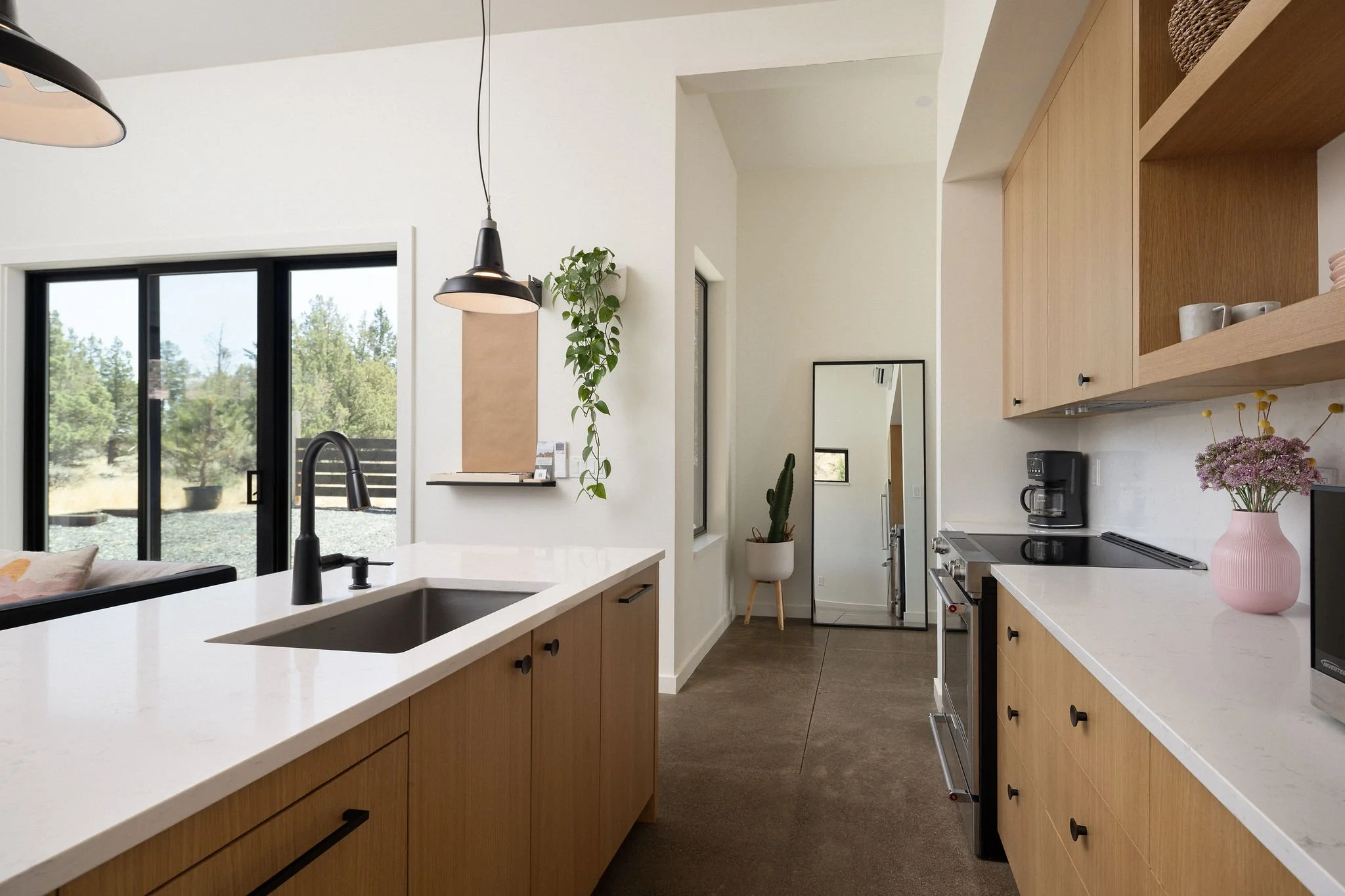 Modern kitchen with wooden cabinets, white countertops, black faucet, black pendant lights, sliding glass door, mirror, potted plants, coffee maker, pink vase with flowers, and a small microwave.