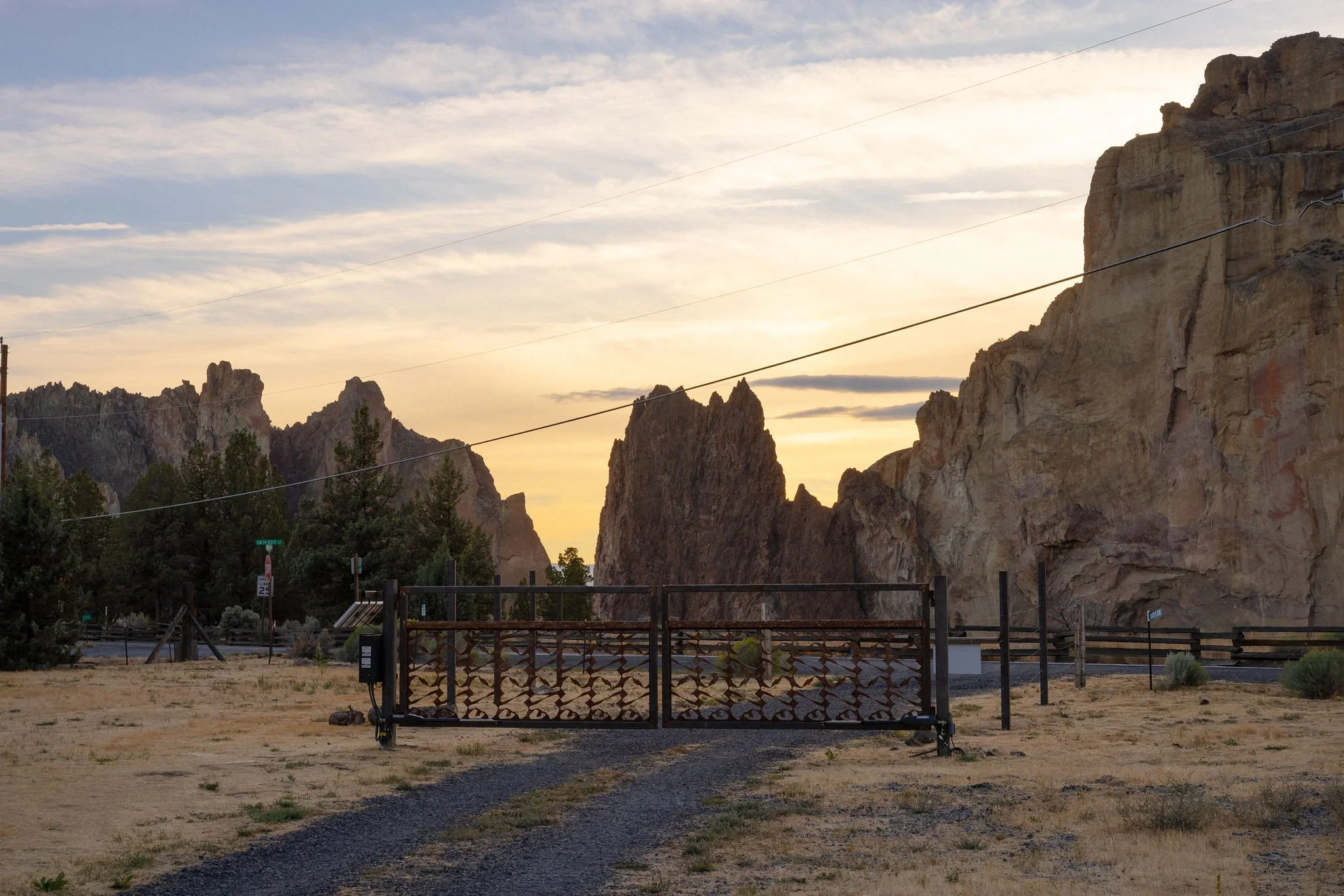 A black decorative gate blocking a dirt path leading towards mountains in the background during sunset, with trees and power lines nearby.