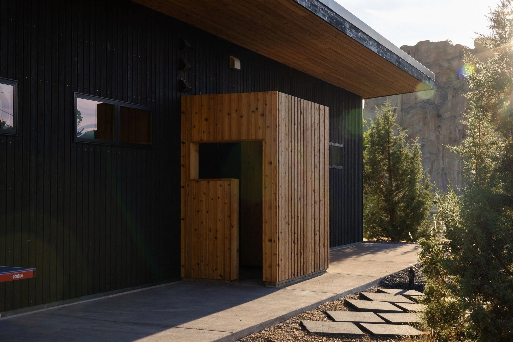 Modern house exterior with black vertical siding and wooden accents, located near trees and rocky cliffs, with sunlight shining on the scene.