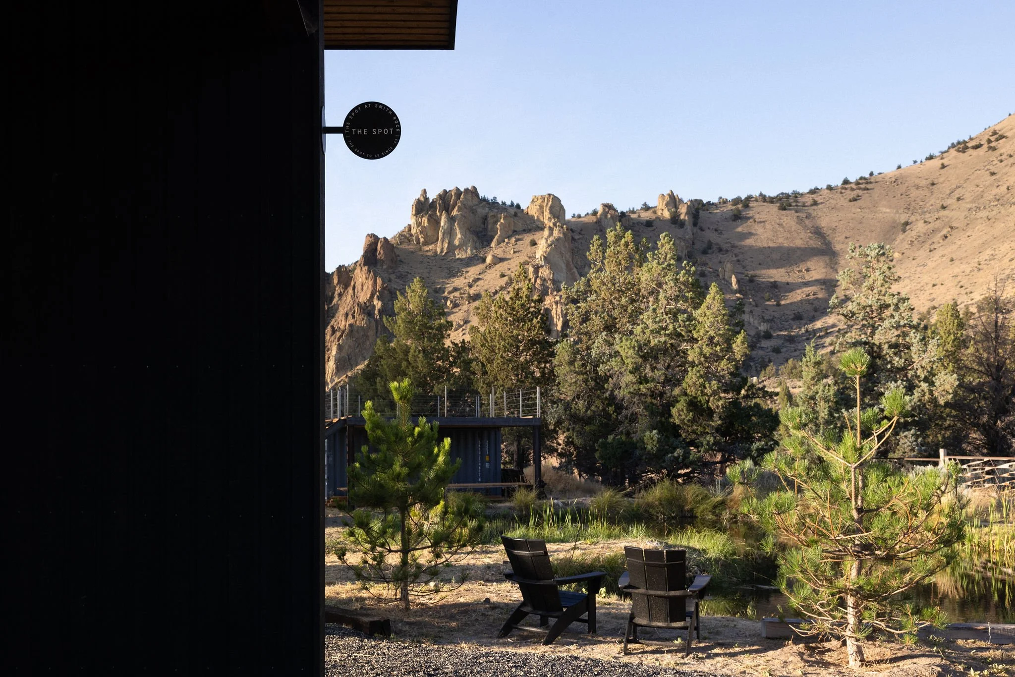 Outdoor scene with two black Adirondack chairs beside a small pond, surrounded by pine trees, with rocky hills and mountains in the background, and part of a dark building visible on the left.