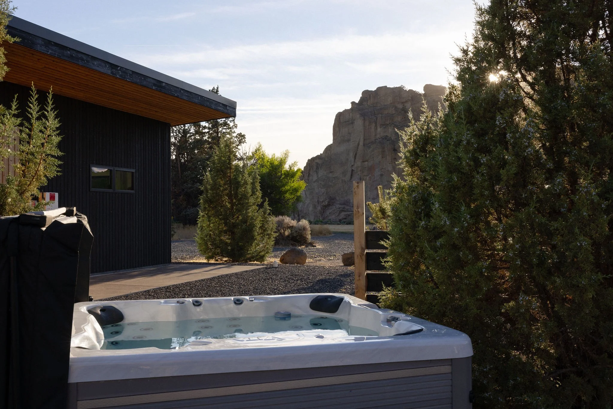 A hot tub on a porch outdoors with trees, rocky landscape, and mountains in the background at sunset.