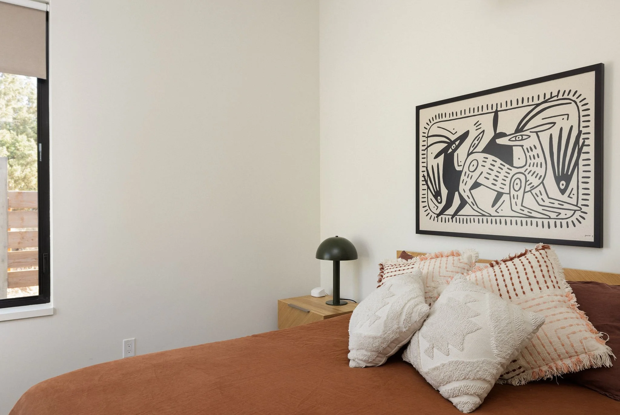 Bedroom with white wall, black framed artwork of abstract animals, wood nightstand, black lamp, white and rust bedding, and pillows.