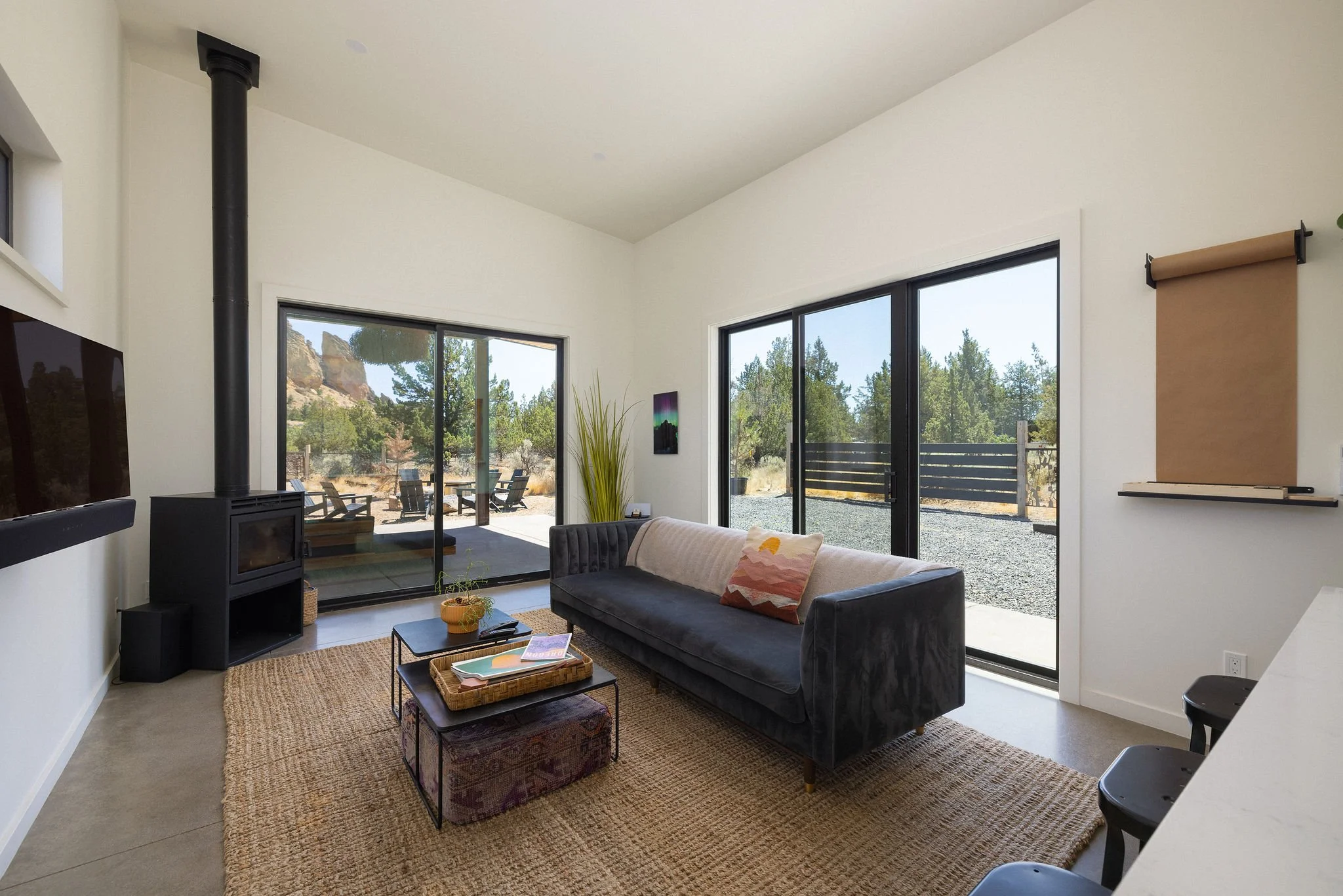 Living room with large sliding glass doors leading outside, a modern black fireplace, a gray velvet sofa with a patterned pillow, a small coffee table with books, a woven rug, and outdoor patio furniture visible through the doors.