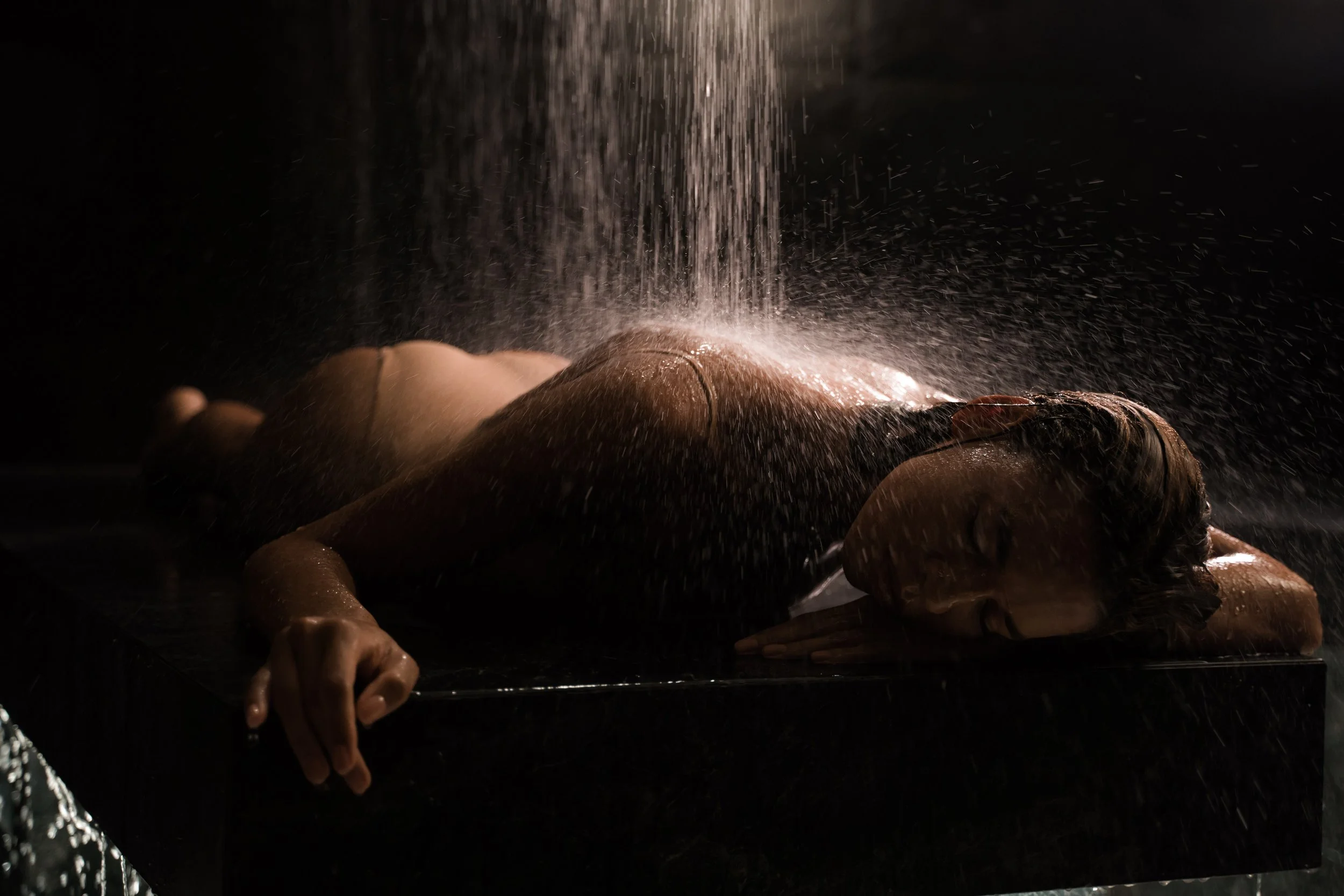 A person lying on a surface with water pouring down on them in a dark setting, creating a dramatic scene with water splashing.