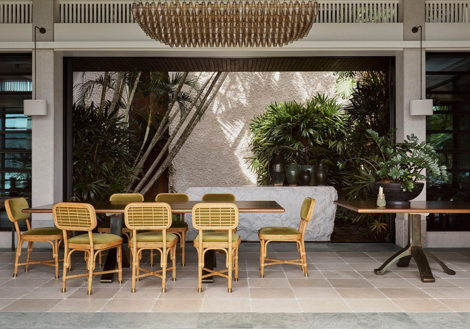 Outdoor patio with a dark wooden dining table and eight yellow rattan chairs with green cushions, surrounded by lush green tropical plants and decorative vases, with a large window and textured wall in the background.