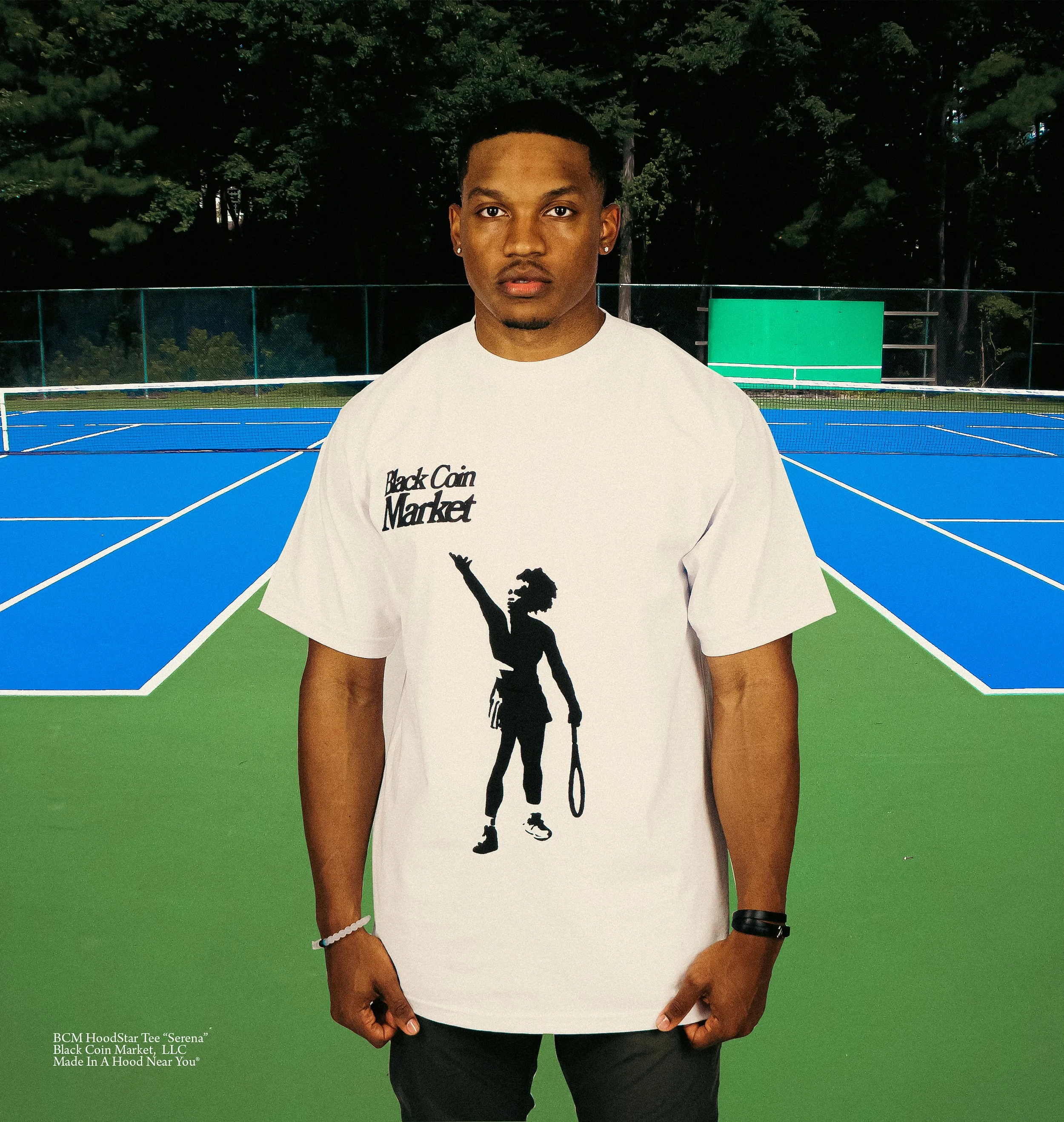 Young man on a tennis court wearing a white BC HoodStar Tee 'Serena' with black silhouette of Serena Williams holding a tennis racket and raising her arm, with a green and blue tennis court background.
