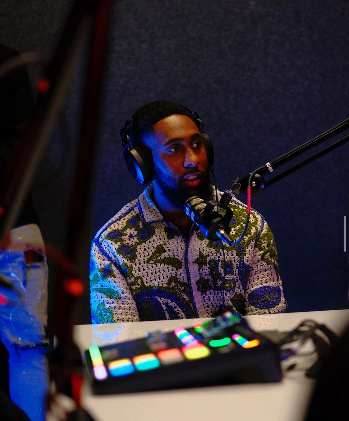 A man with dark skin, black hair, and beard wearing headphones and a patterned shirt, speaking into a professional microphone in a recording studio.