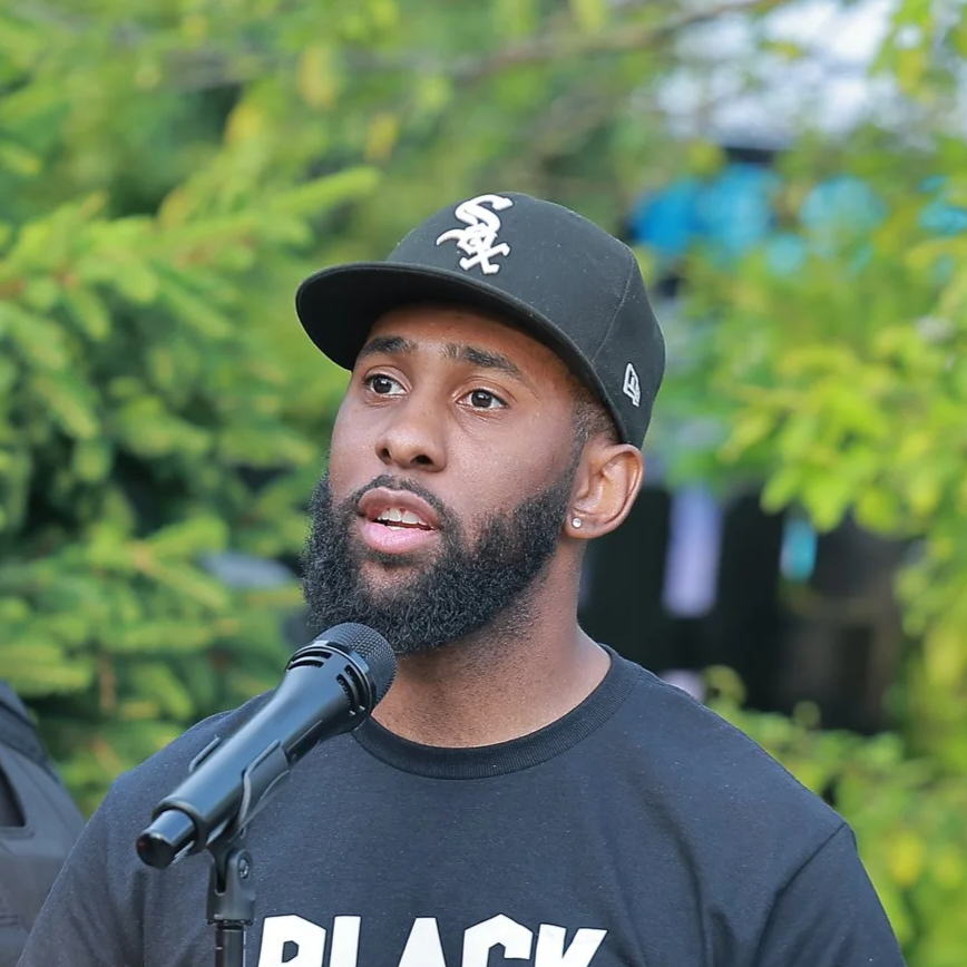 A man with a beard and earrings speaking into a microphone outdoors, wearing a black cap and a black T-shirt with white text.