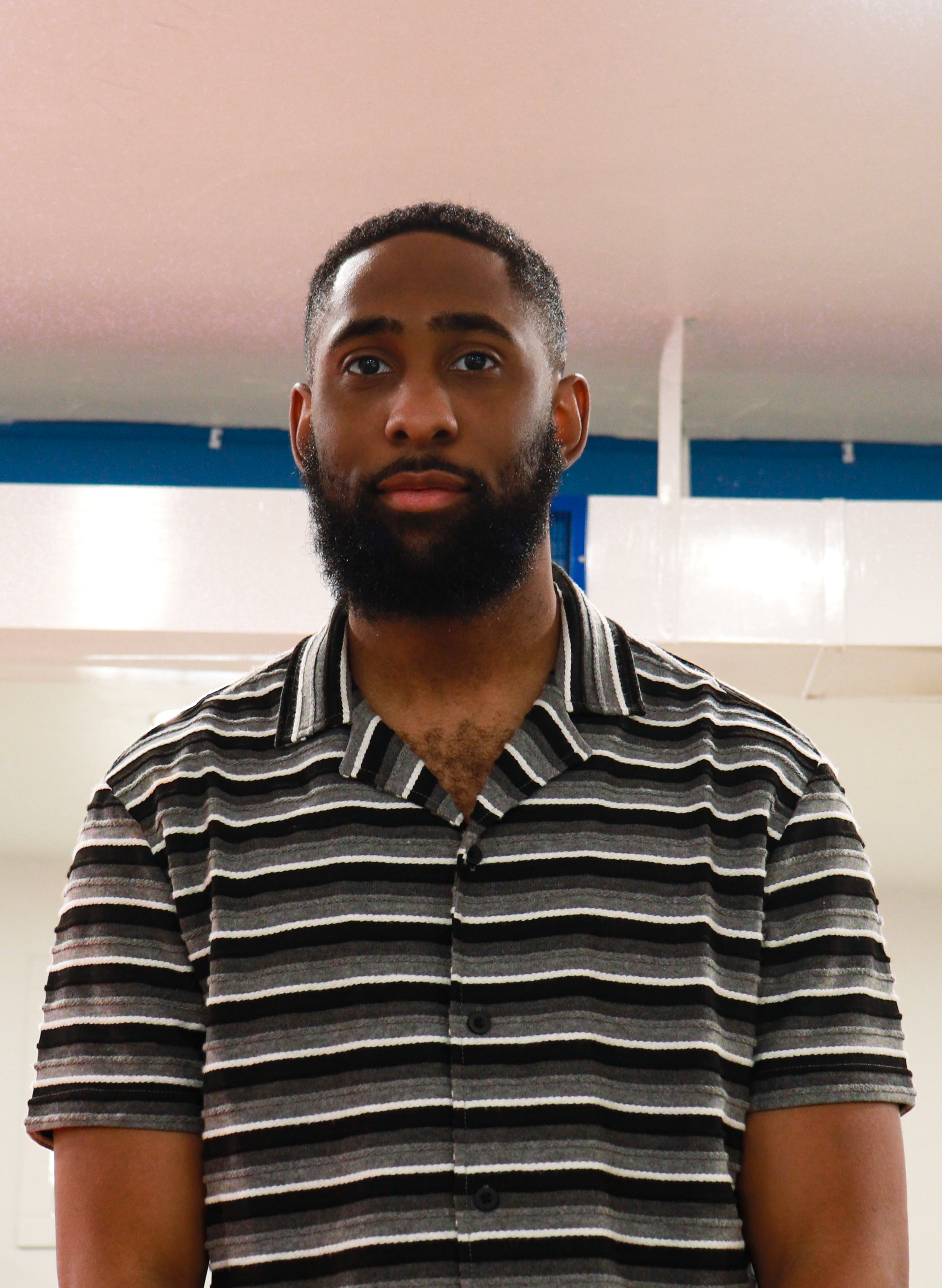 A young man with a beard and short hair wearing a black, gray, and white striped short-sleeve shirt standing indoors.