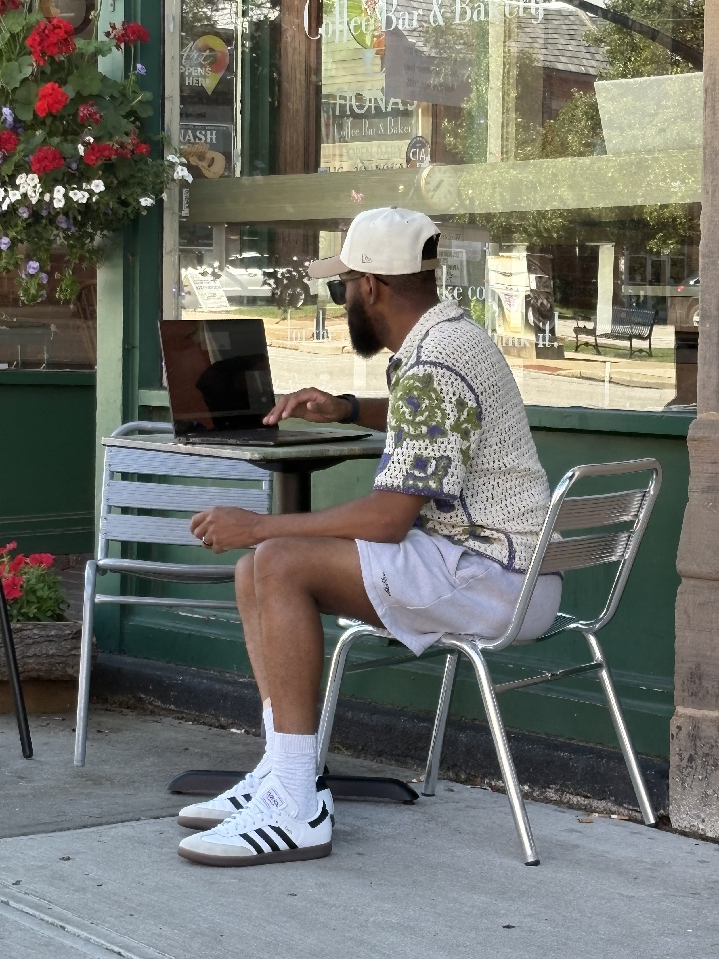 Man sitting outside at a cafe, working on a laptop, wearing a white cap, sunglasses, patterned shirt, grey shorts, white Adidas sneakers, and white socks, with a green storefront and flower basket in the background.