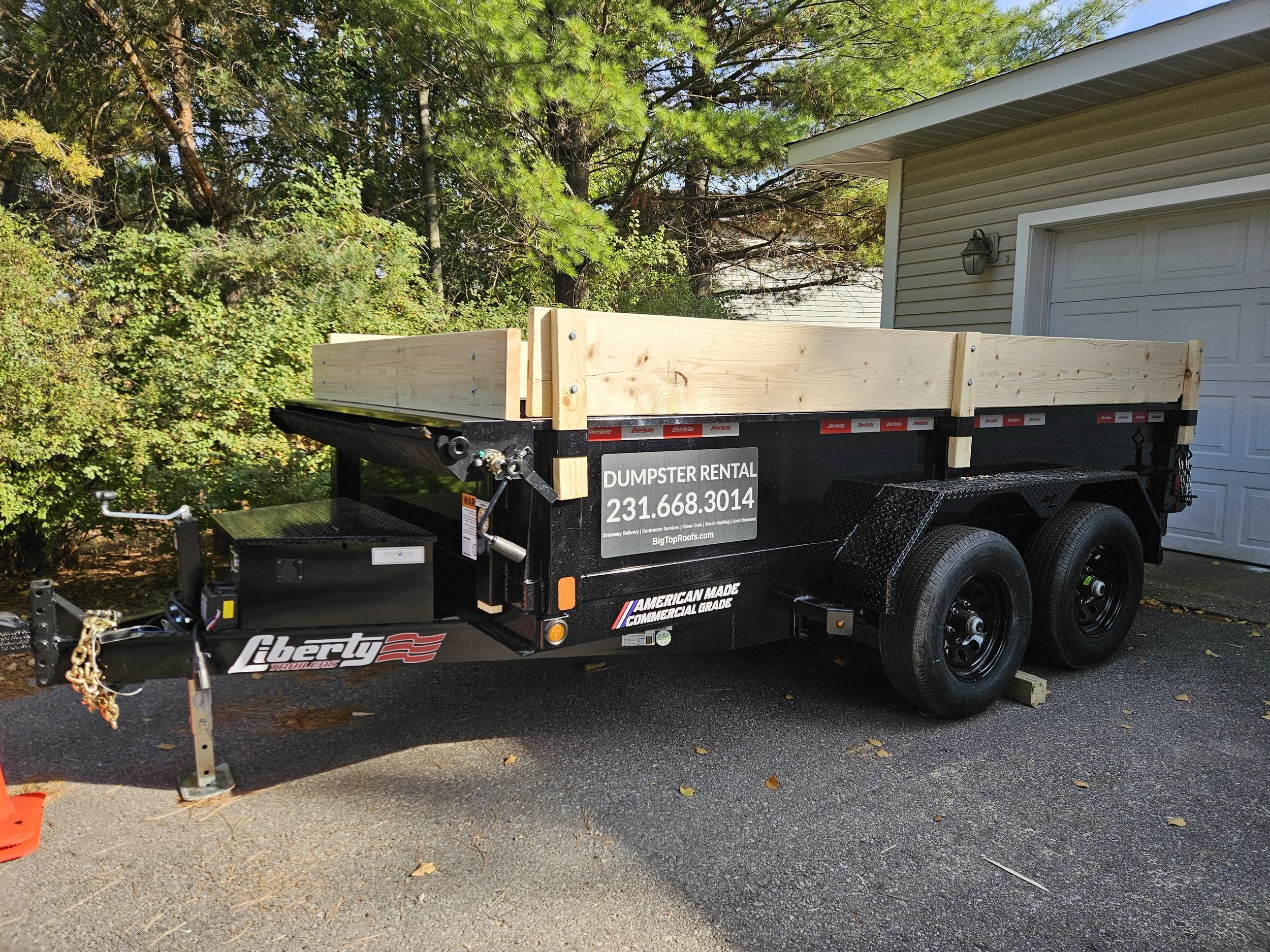 Black utility trailer with double axles and wooden rails, parked in front of a garage, surrounded by trees. It features "Dumpster Rental" and a contact number on the side.