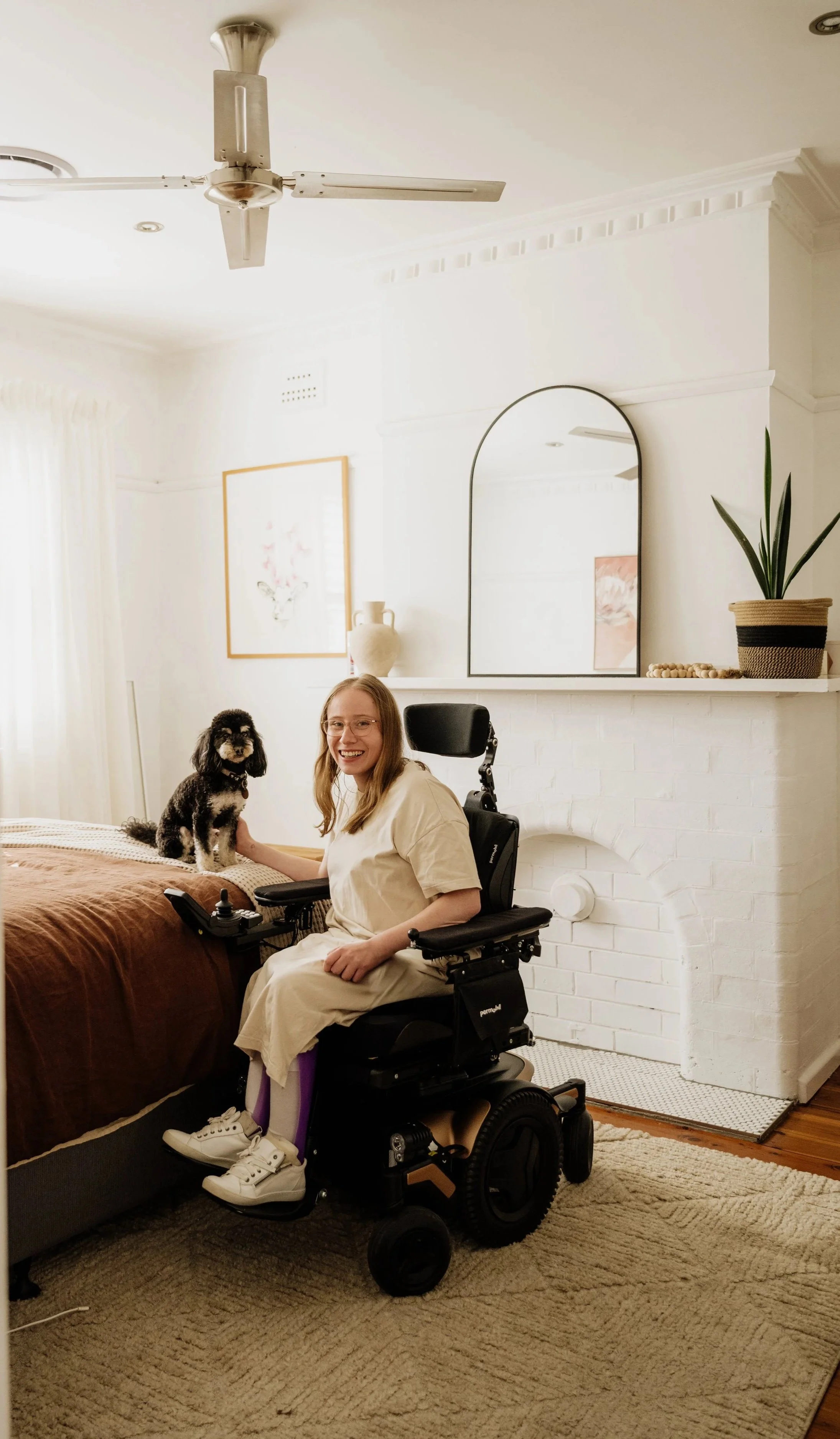 Elle smiling in her wheelchair beside a bed, gently holding a small dog in a bright, cosy bedroom.