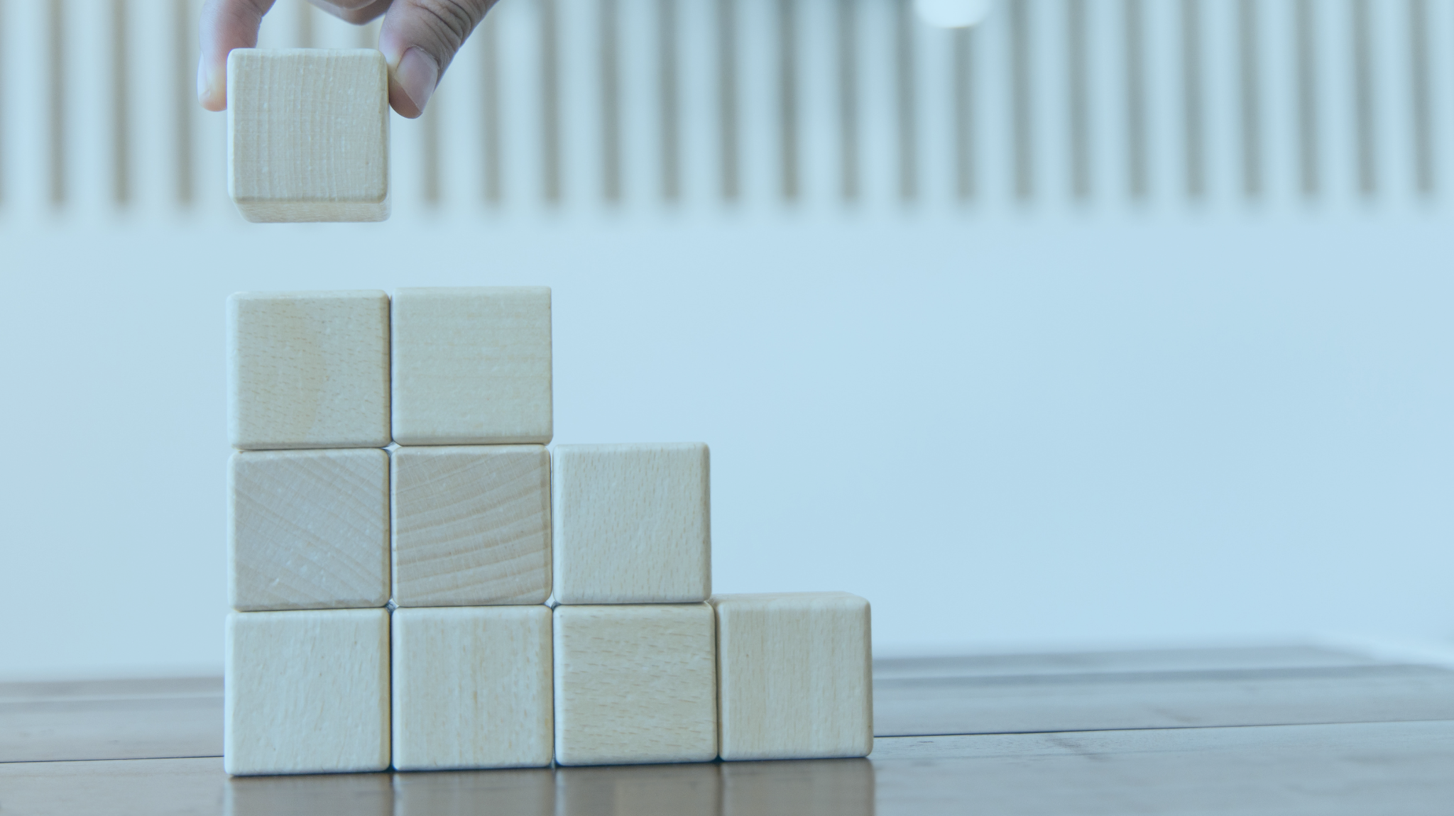 A hand stacking a wooden cube on top of a pyramid of similar cubes on a wooden surface.