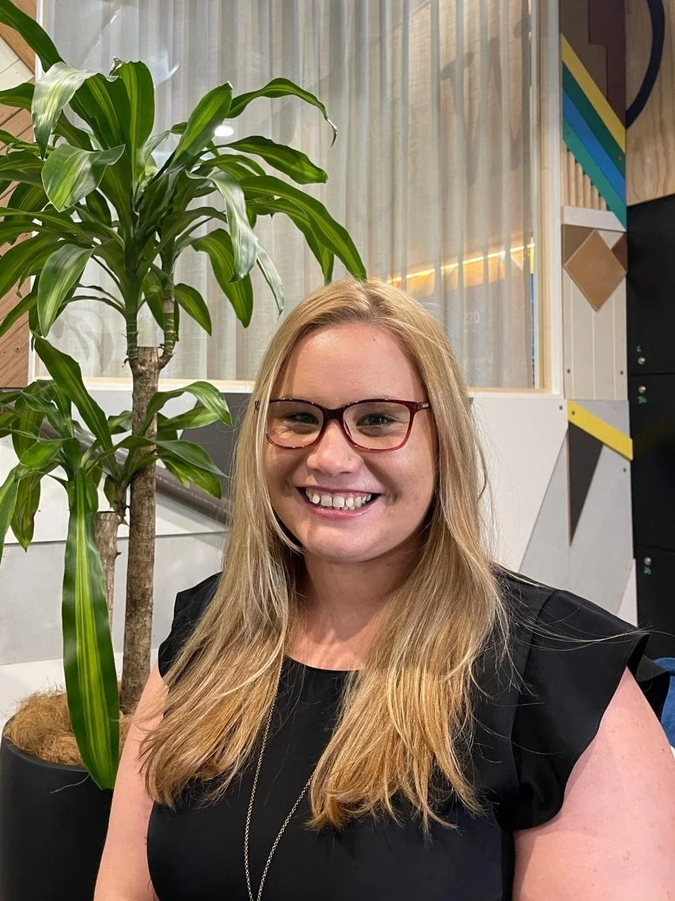 A woman with blonde hair wearing glasses and a black top, smiling, sitting in front of a large potted plant with long green leaves in an indoor setting.