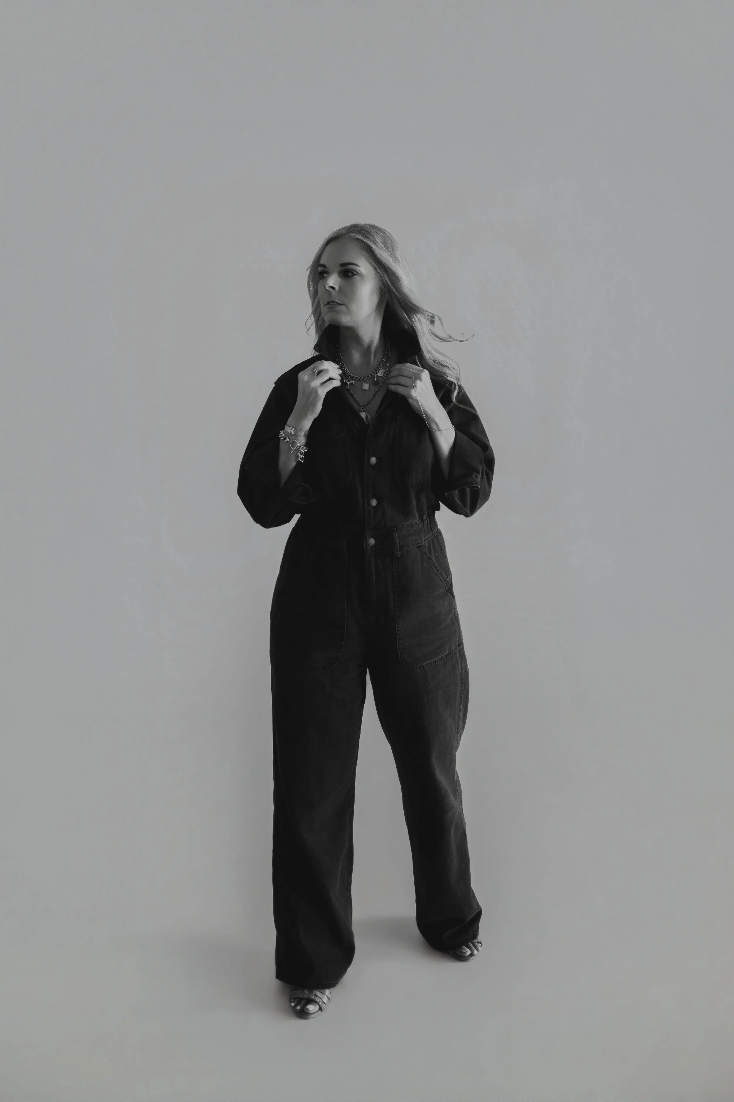 Black and white photo of a woman with wavy hair wearing a dark jumpsuit and jewelry, standing against a plain backdrop.