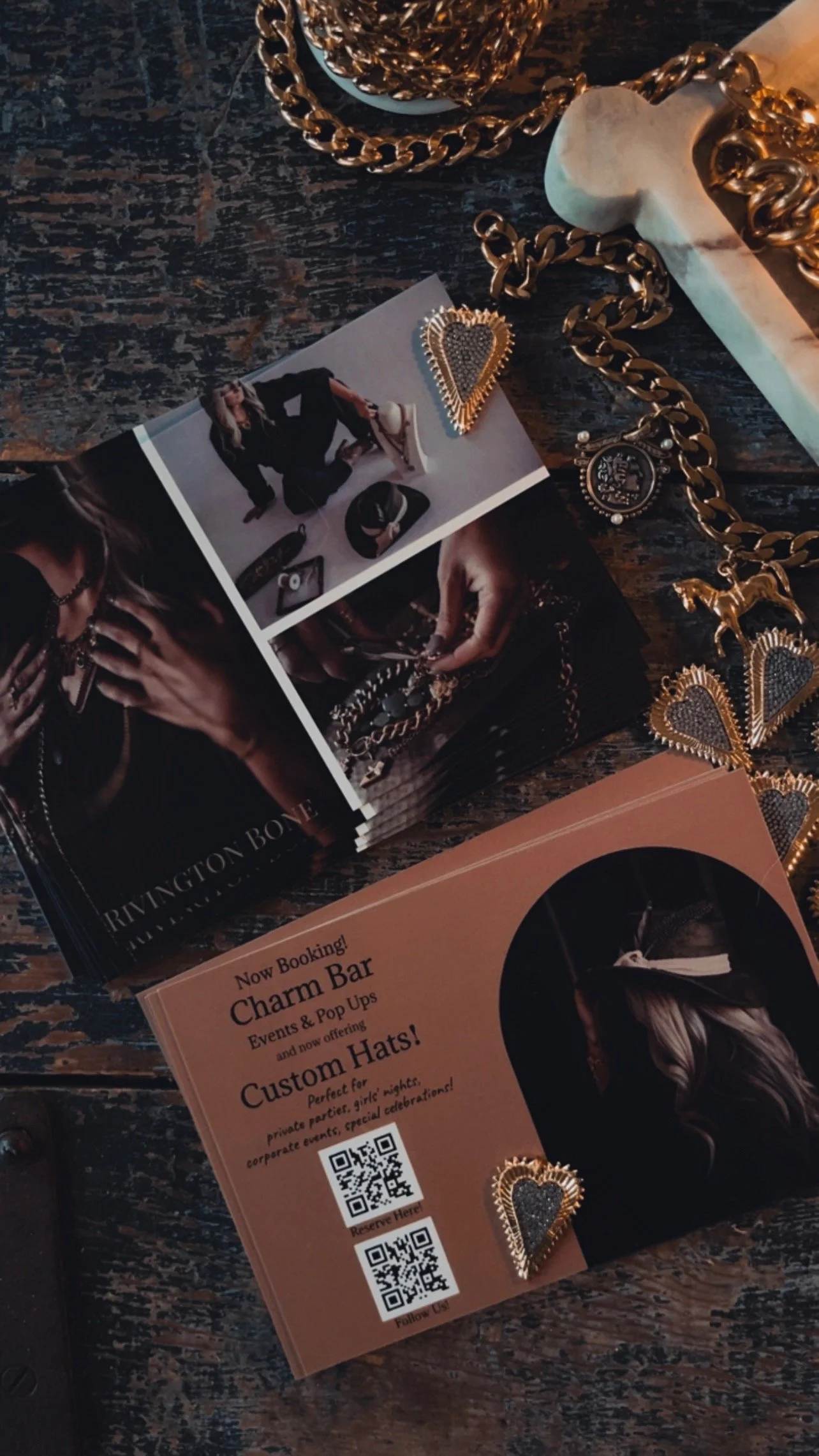 A promotional flyer for Charm Bar with QR codes, jewelry pieces including earrings, chains, and a heart-shaped pendant, and a brochure featuring models wearing jewelry, all on a rustic wooden surface.