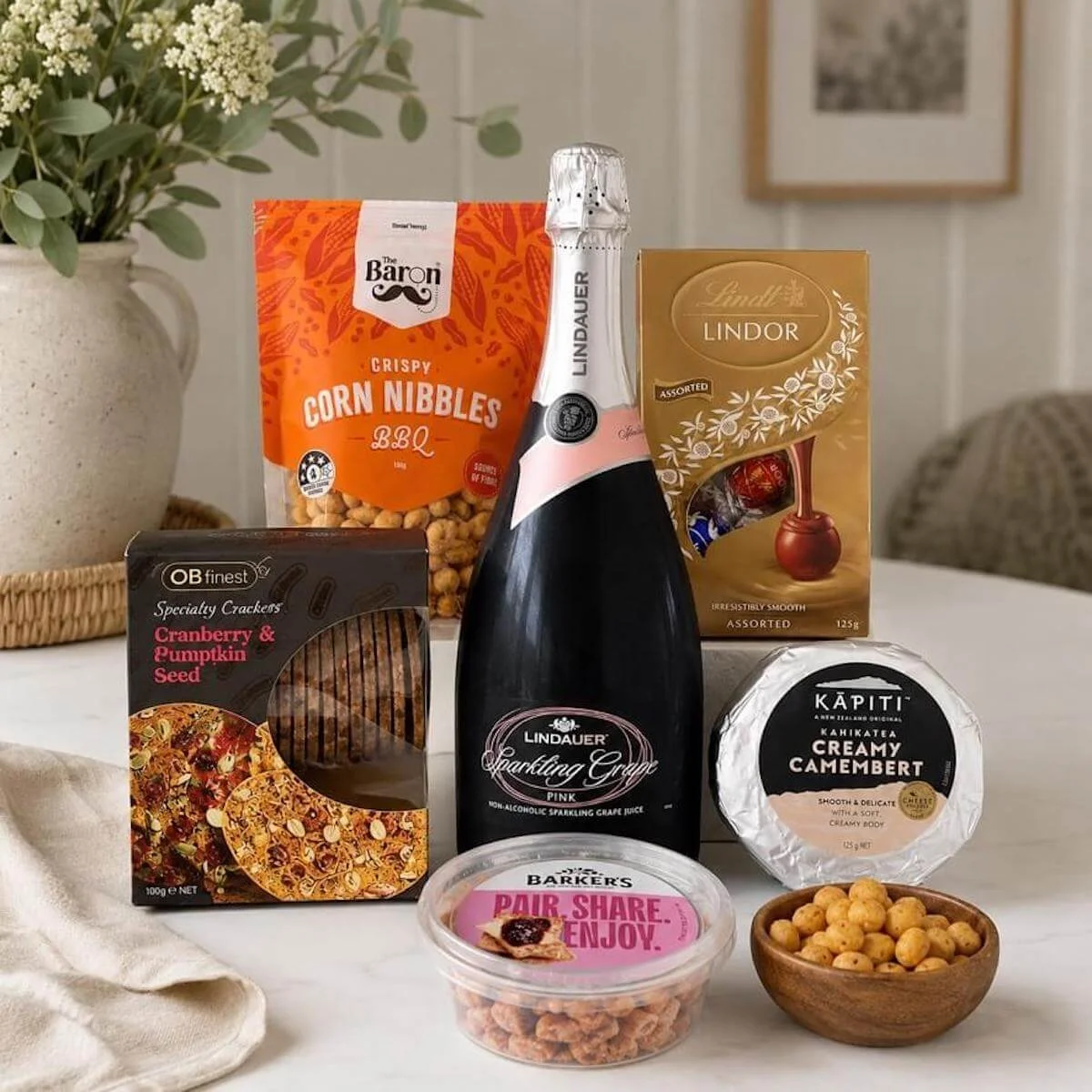 Gift hamper items displayed on a table. Bottle of grape juice, cheese, fruit paste, chocolates, corn nibbles and crackers.