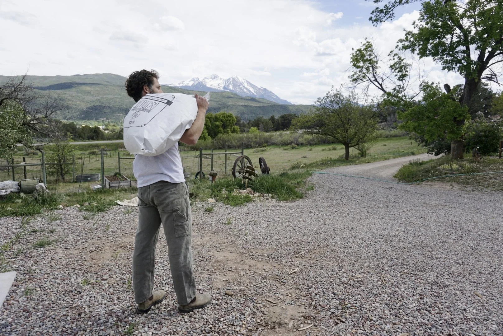 A man carrying a 50lb bag of Roaring Fork Mill flour over his shoulder, standing outdoors in a rural area with trees, a gravel path, and green hills with snow-capped mountains in the background.