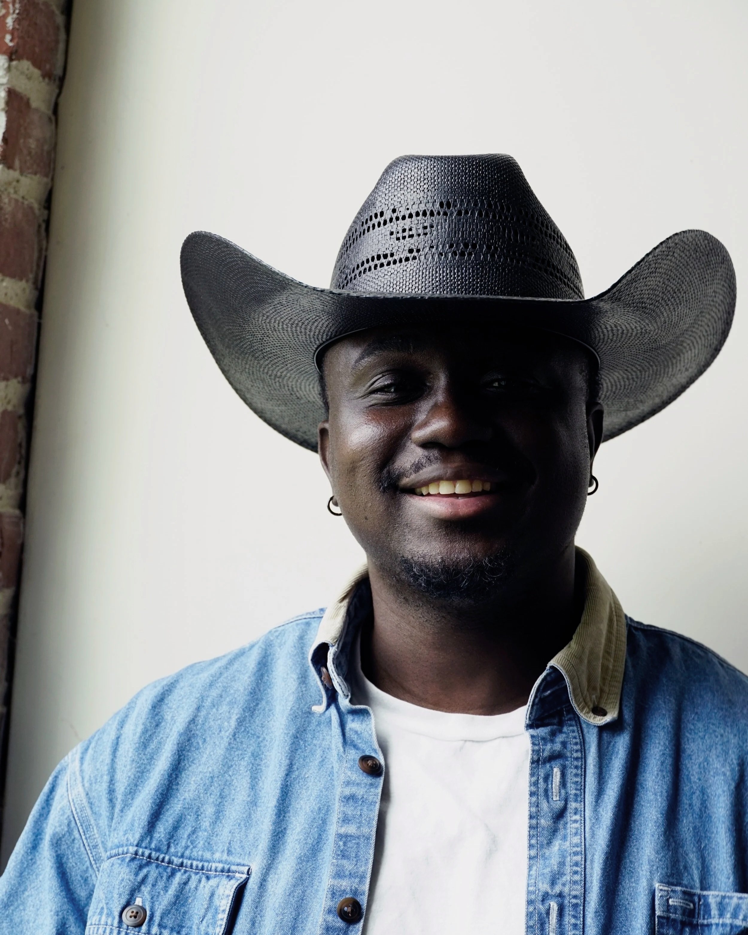 A man wearing a cowboy hat, a denim jacket, and a white T-shirt, smiling in front of a plain background with a brick wall on the left.