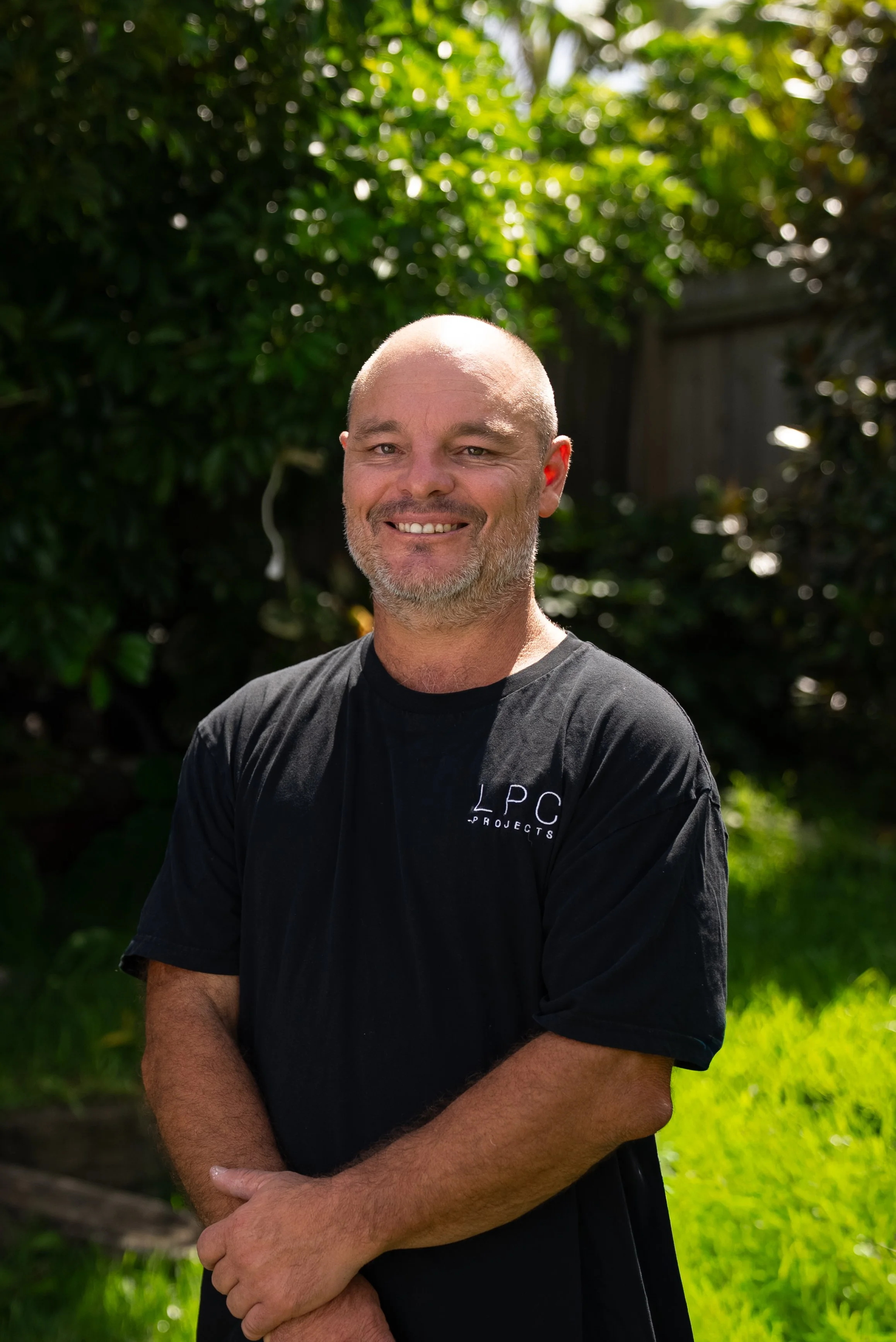 A smiling man with a beard, wearing a navy blue work shirt with 'L.P.C Projects' logo, standing inside a construction or renovation site.