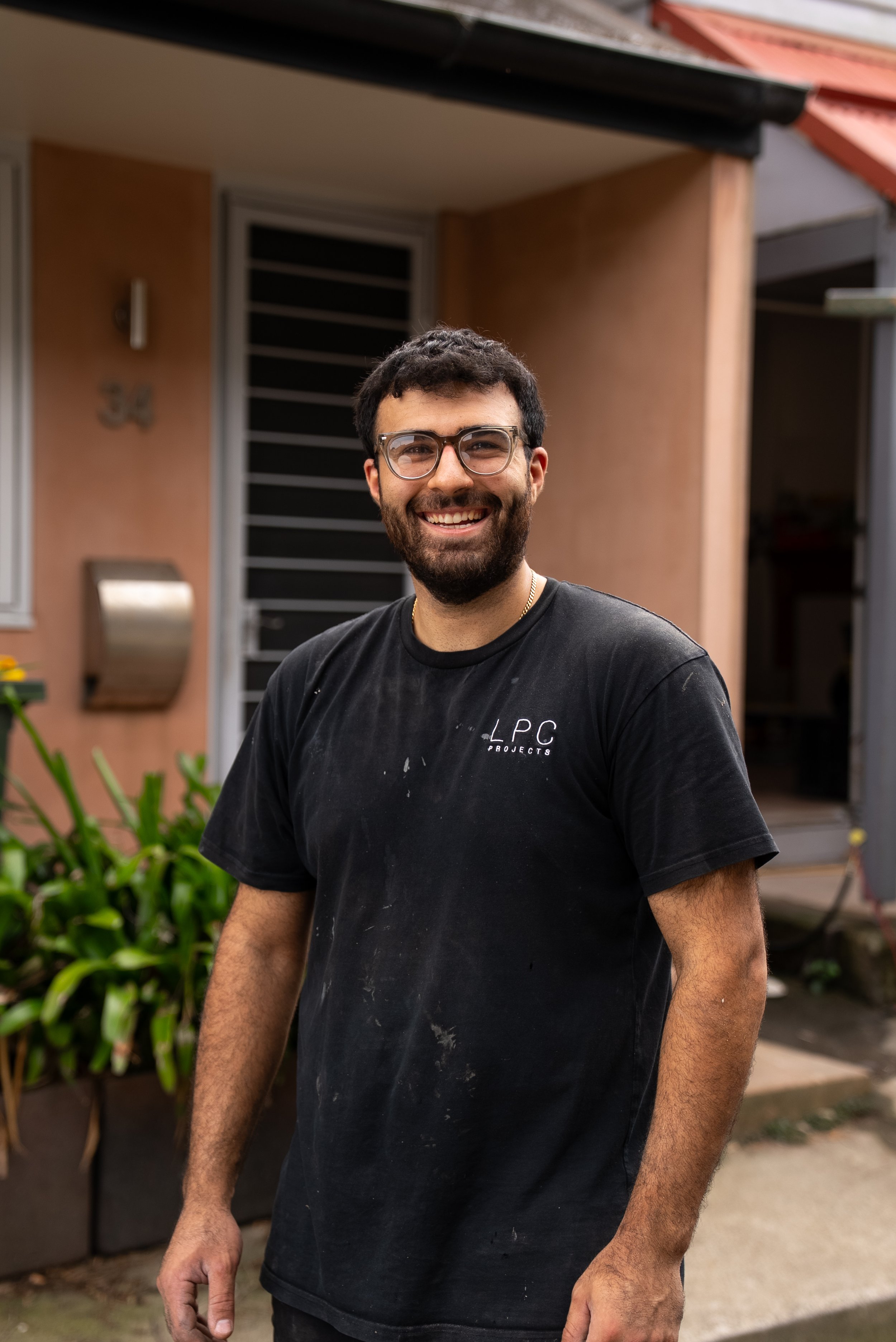 A young man with short brown hair and a beard, wearing a black t-shirt with white text, standing outdoors in front of green trees.