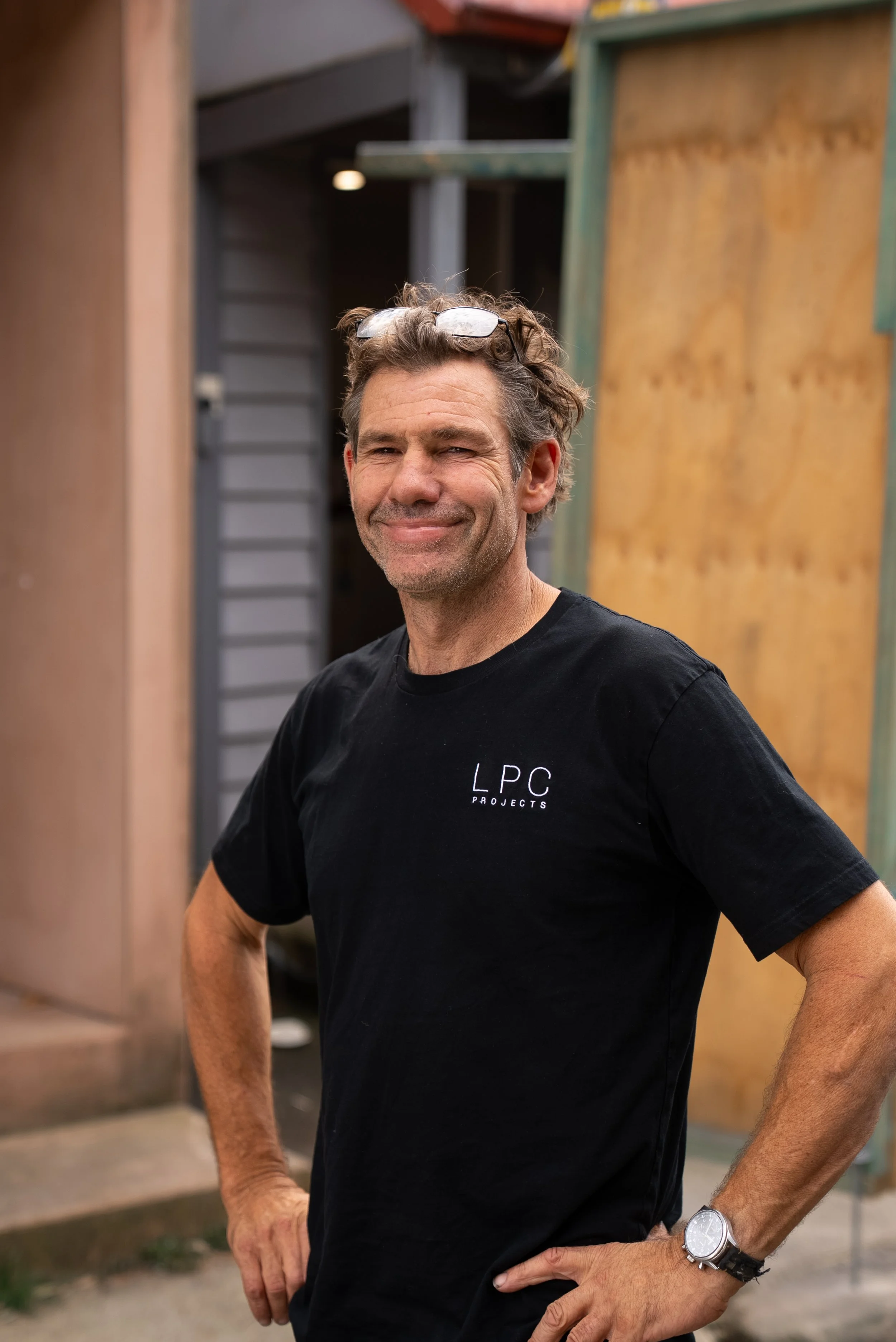 A young man with light brown hair, blue eyes, and a slight smile standing outdoors in a wooded area, wearing a black t-shirt with 'LPC PROJECTS' written on it.