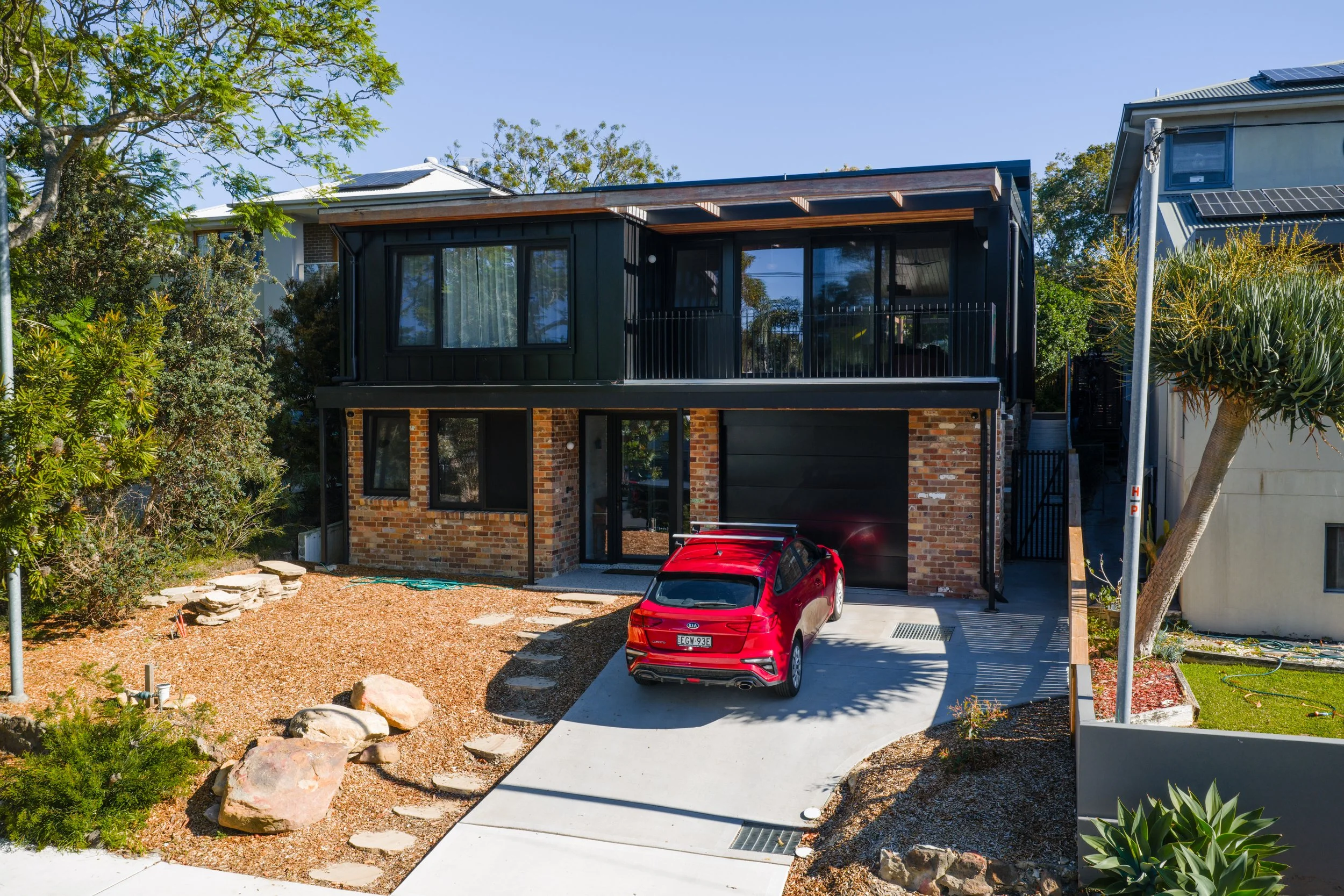 Living room with beige armchairs, a round marble coffee table with vases and dried flowers, a white TV stand with a flat-screen TV, a tall floor lamp, and an open kitchen with white cabinets and bar stools.