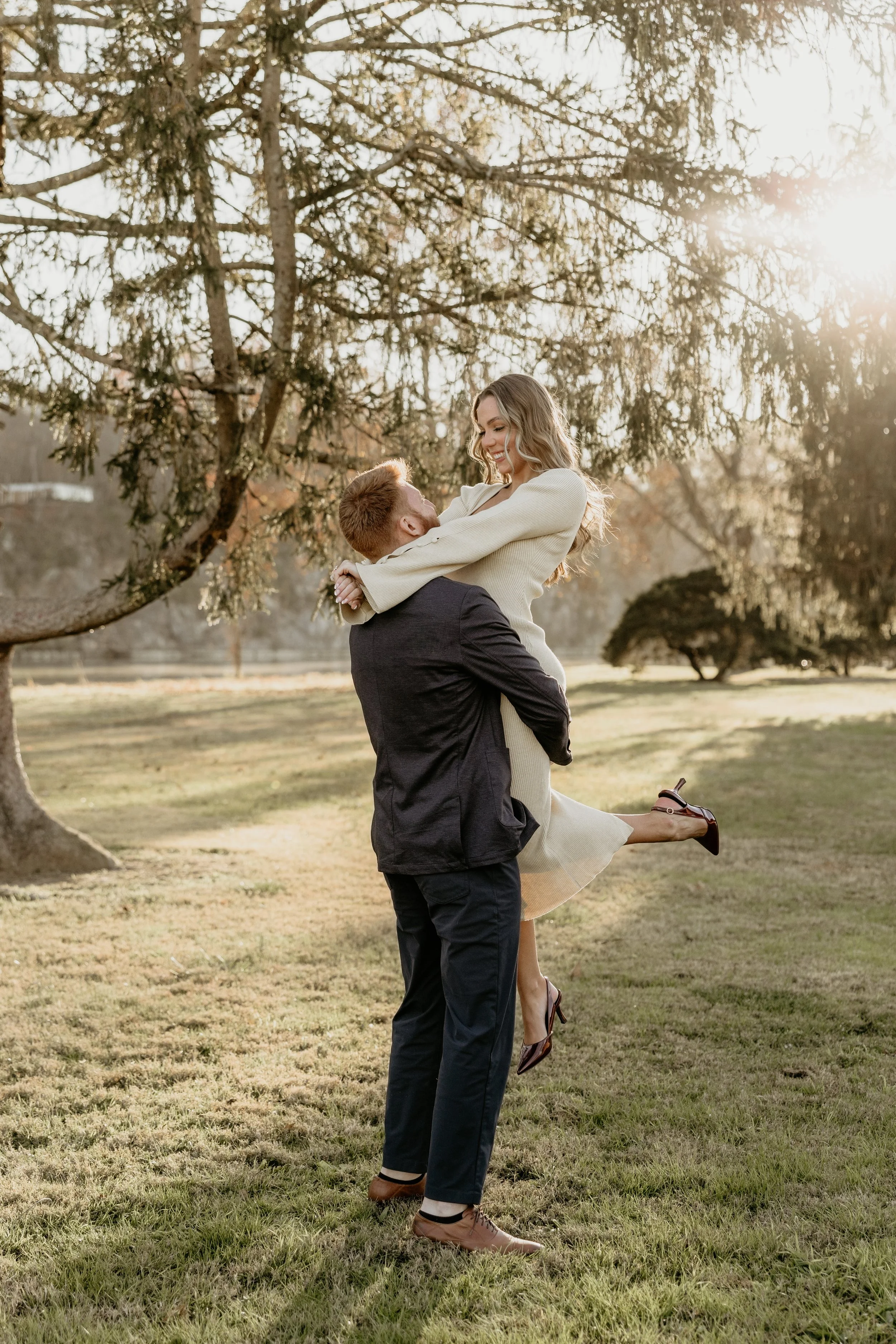 A man lifting a woman in a field during sunset, with large trees in the background, both smiling and embracing each other.