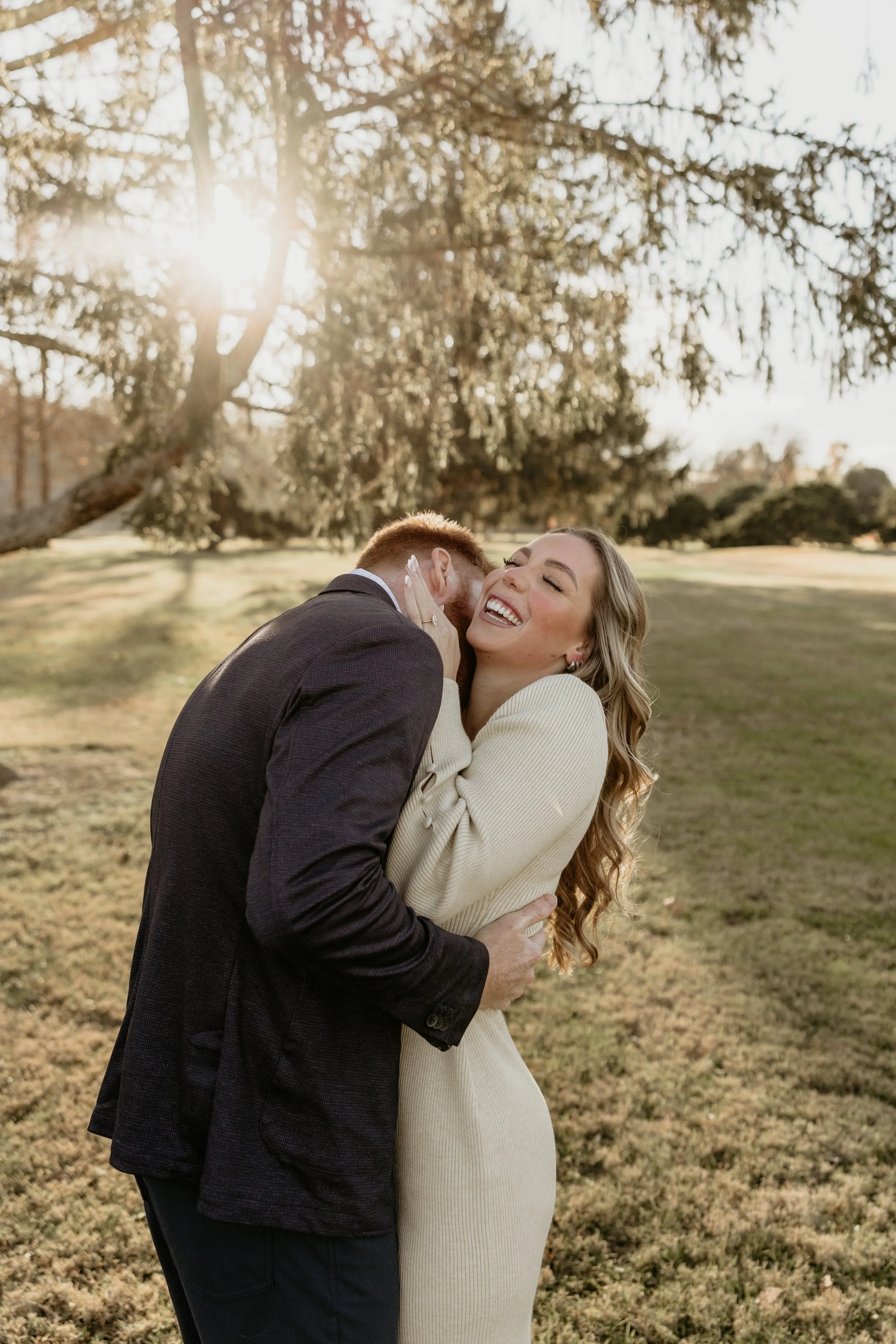 A man and woman sharing a joyful moment outdoors, with the man kissing the woman's cheek as she laughs. They are embracing in a grassy area with trees and sunlight filtering through the branches.