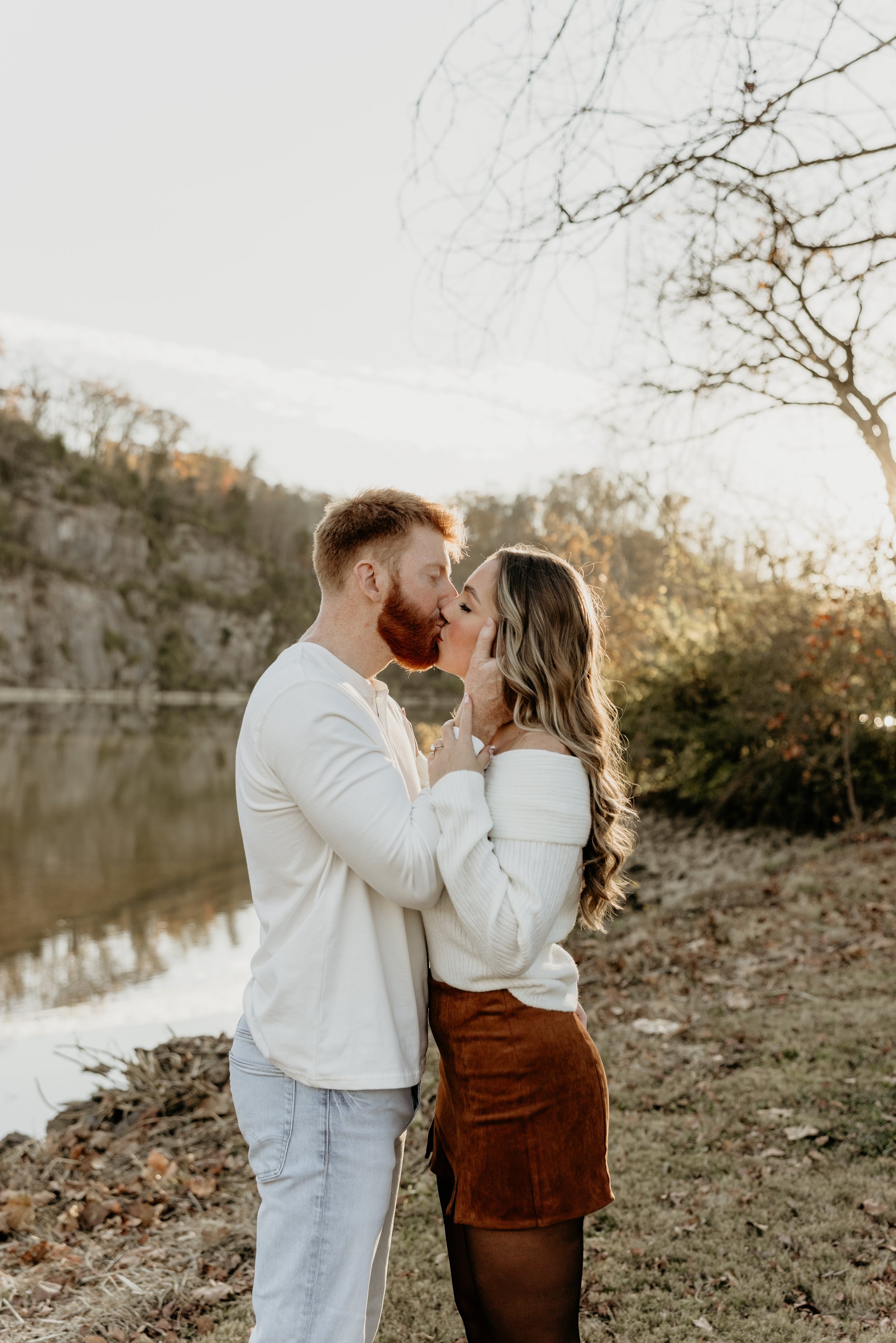A couple kissing outdoors by a river at sunset with trees in the background.