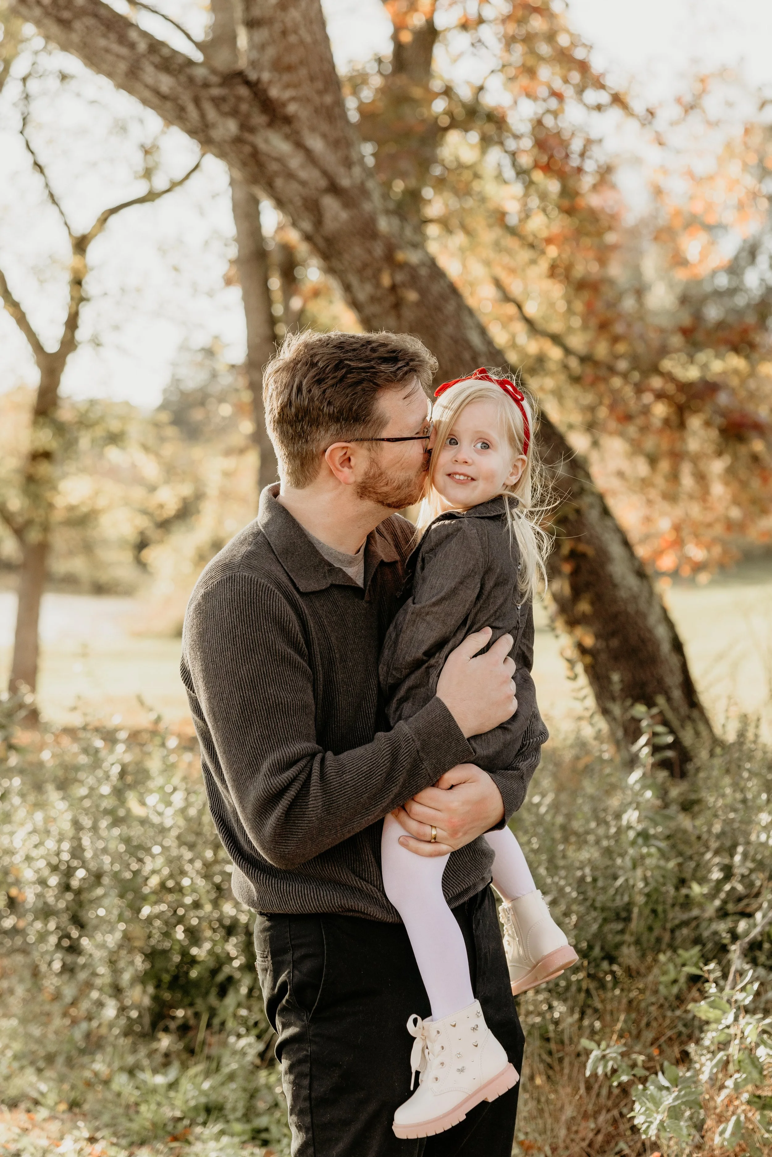 A man holding a young girl outdoors in a park during autumn, with trees in the background with fall foliage.