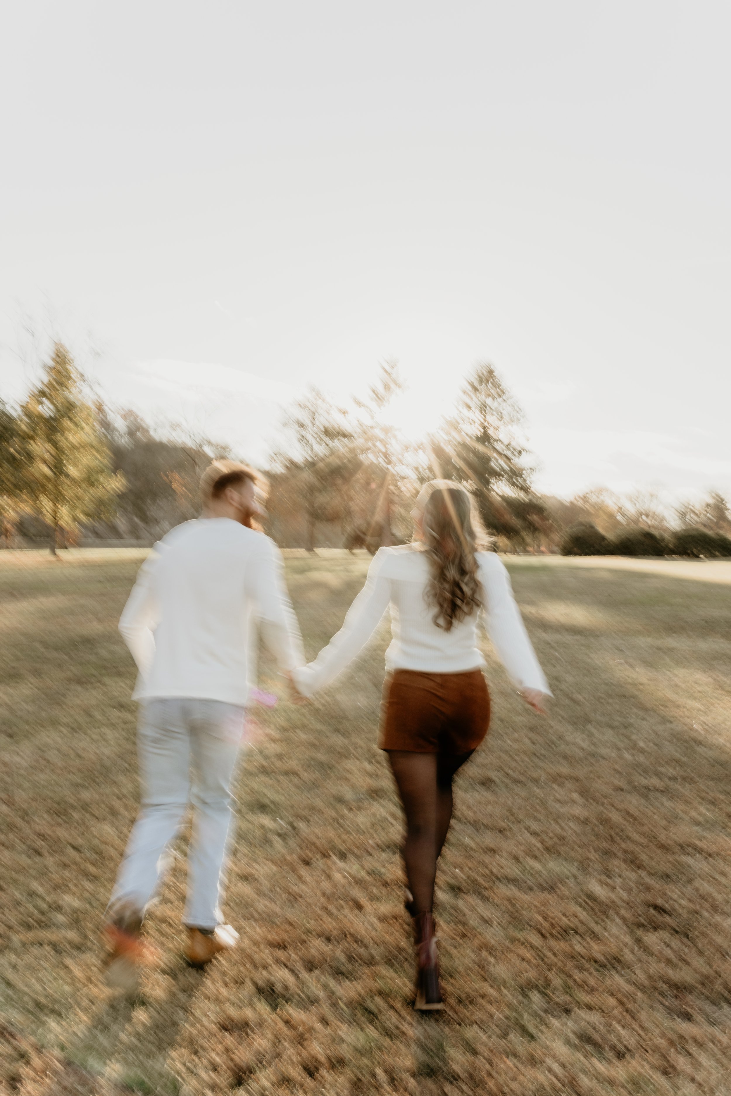 A couple running hand in hand through an open field with trees and the setting sun in the background, captured in warm, soft sunlight.