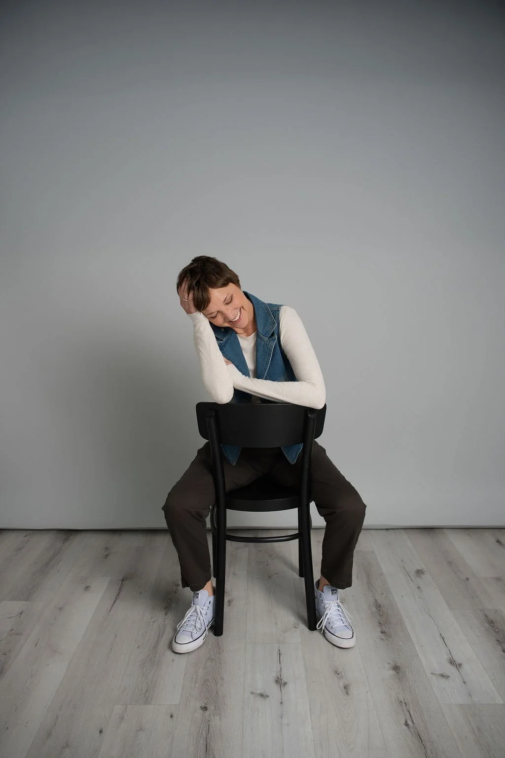 A woman sitting on a black chair, leaning forward with a big smile, head resting on her hand, against a plain gray background. She is wearing a white long-sleeve shirt, a blue vest, brown pants, and white sneakers.