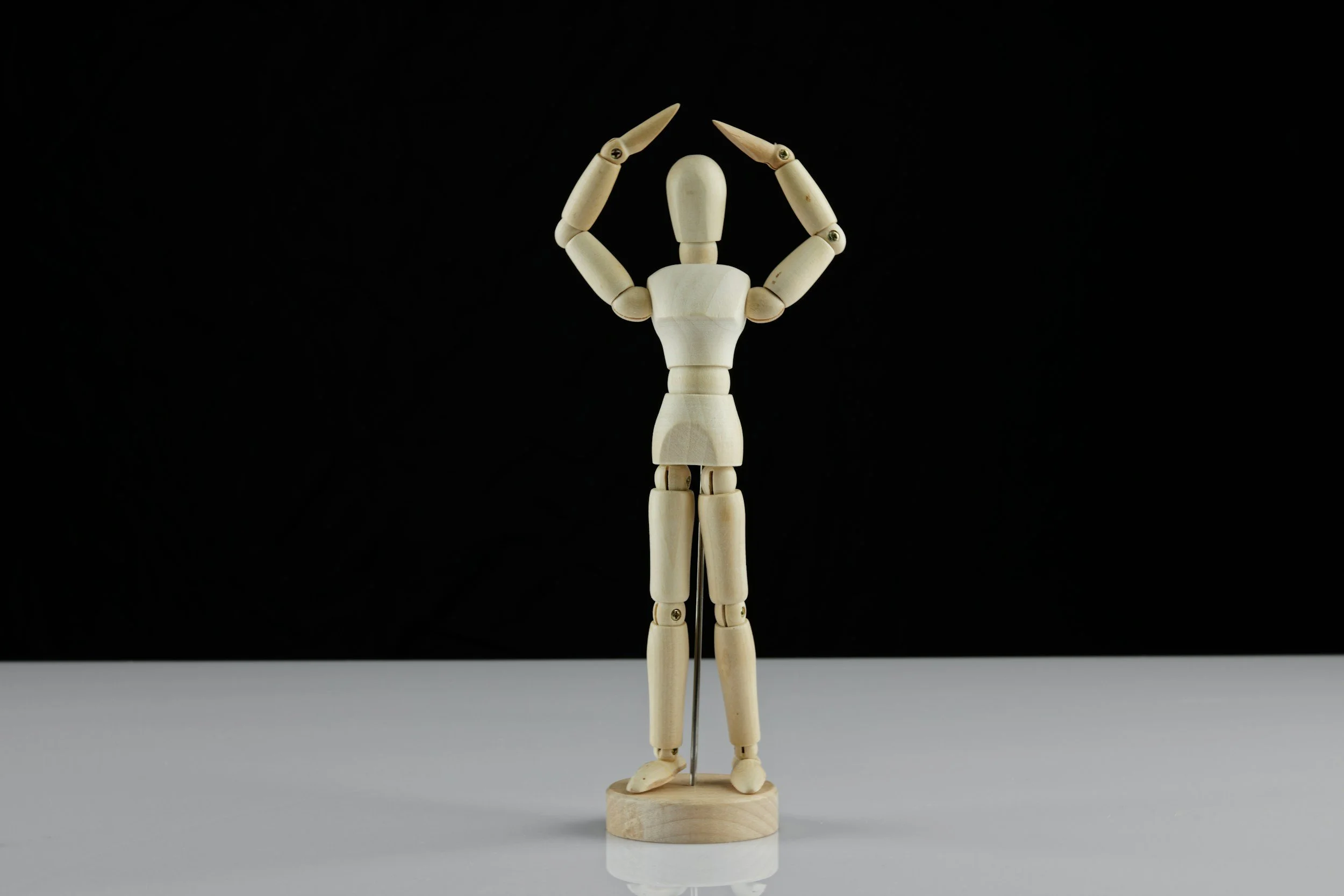 A wooden artist mannequin with arms raised forming a heart shape against a black background.