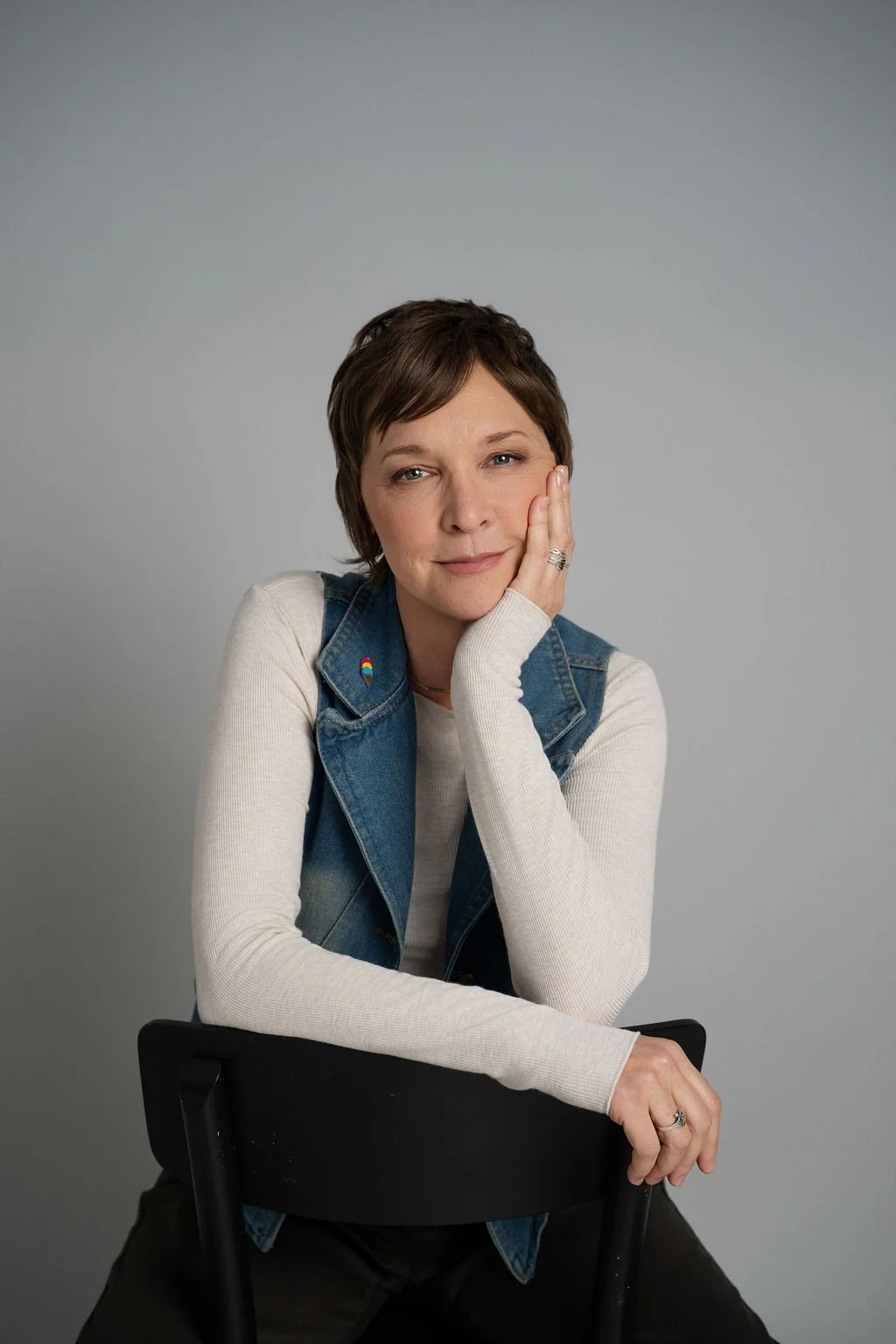 A woman with short brown hair, wearing a denim vest and cream long-sleeve top, sitting on a black chair in front of a gray background, resting her head on her hand and smiling softly.