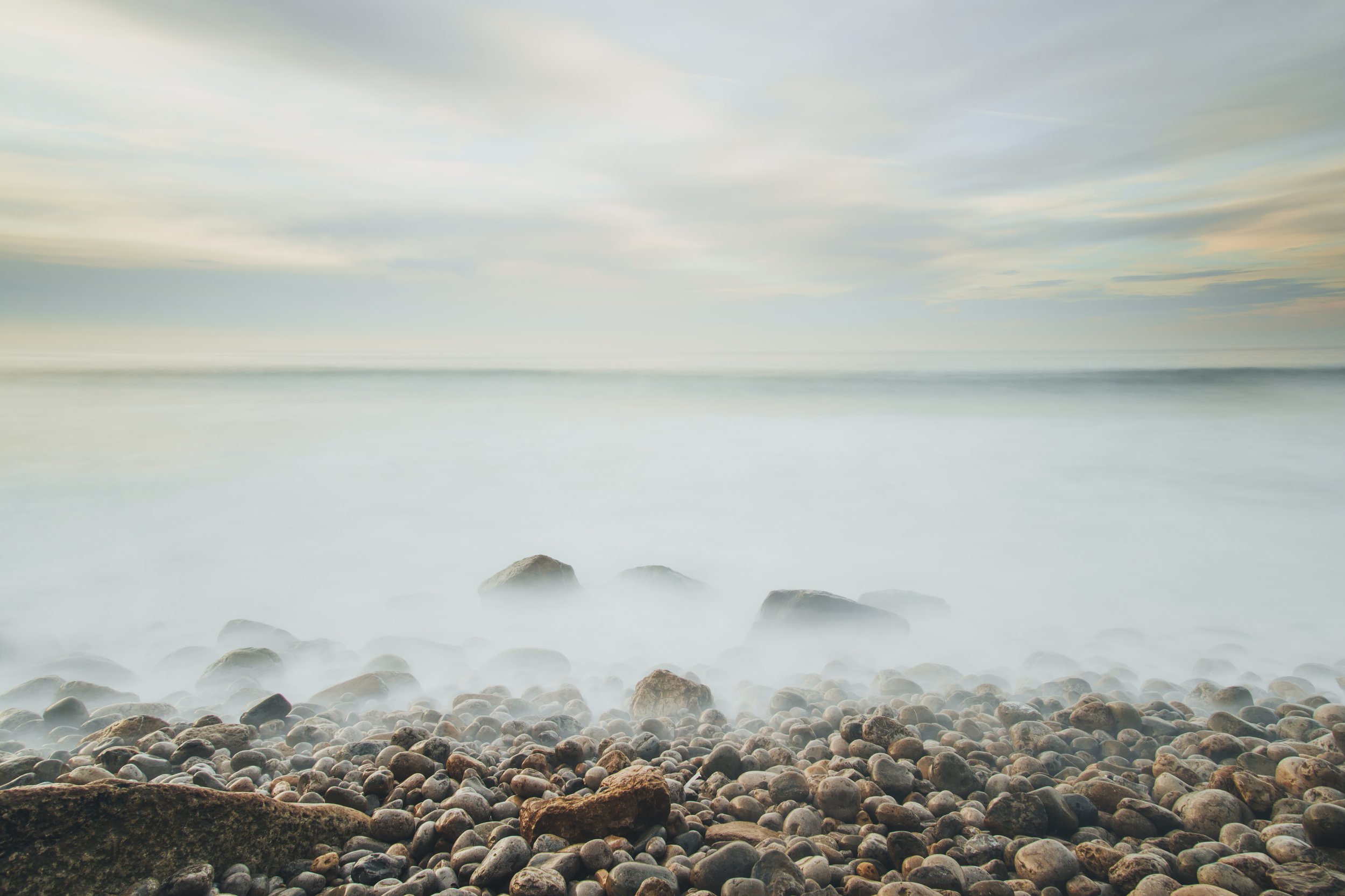 A rocky beach with smooth pebbles in the foreground and calm water with gentle waves in the background under a cloudy sky.
