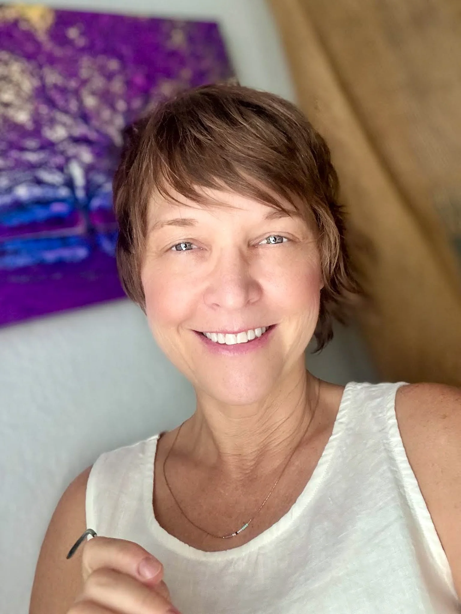 Smiling woman with short brown hair and blue eyes taking a selfie indoors, with a colorful painting in the background.