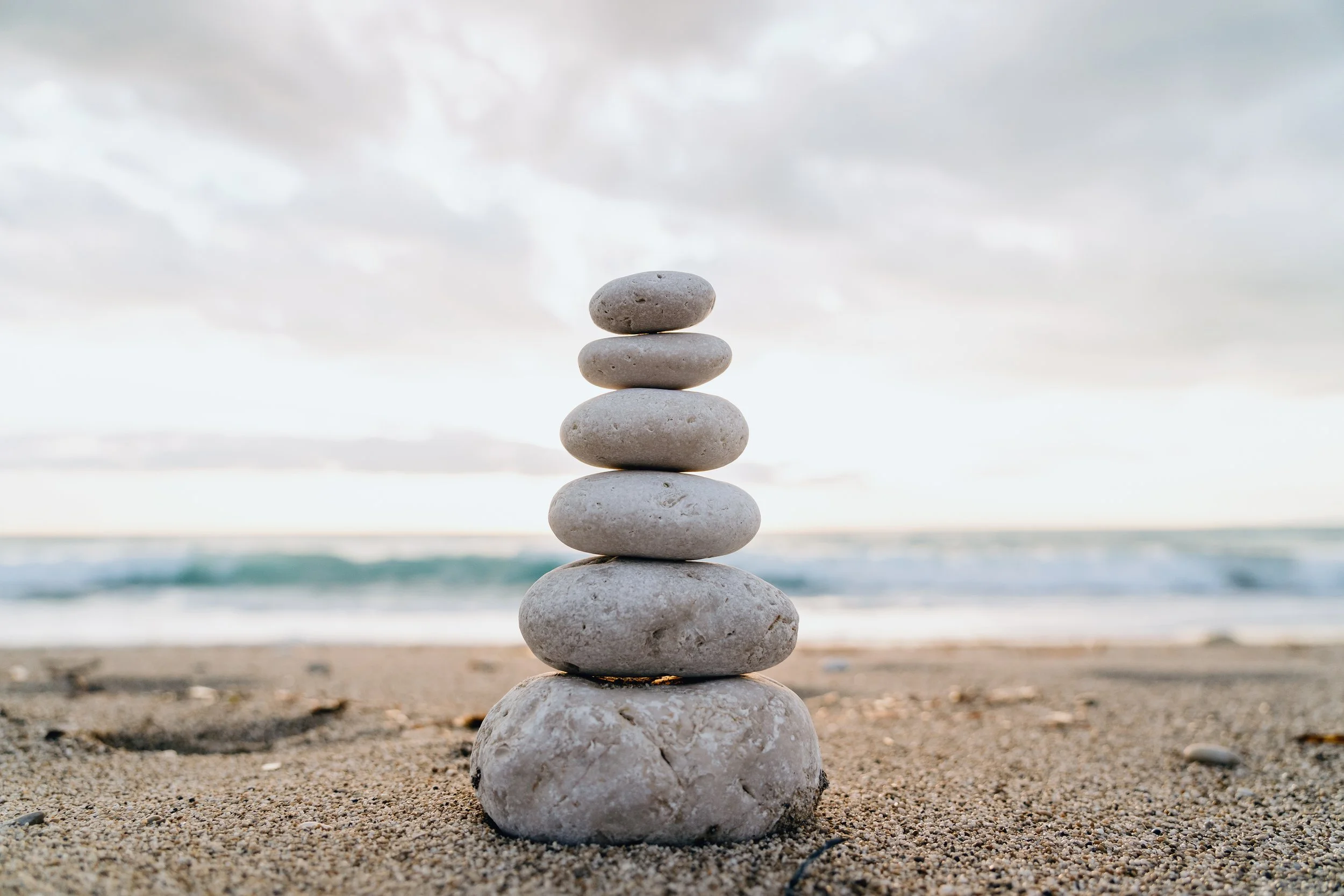Stacked smooth gray stones on sandy beach with ocean and cloudy sky in background.
